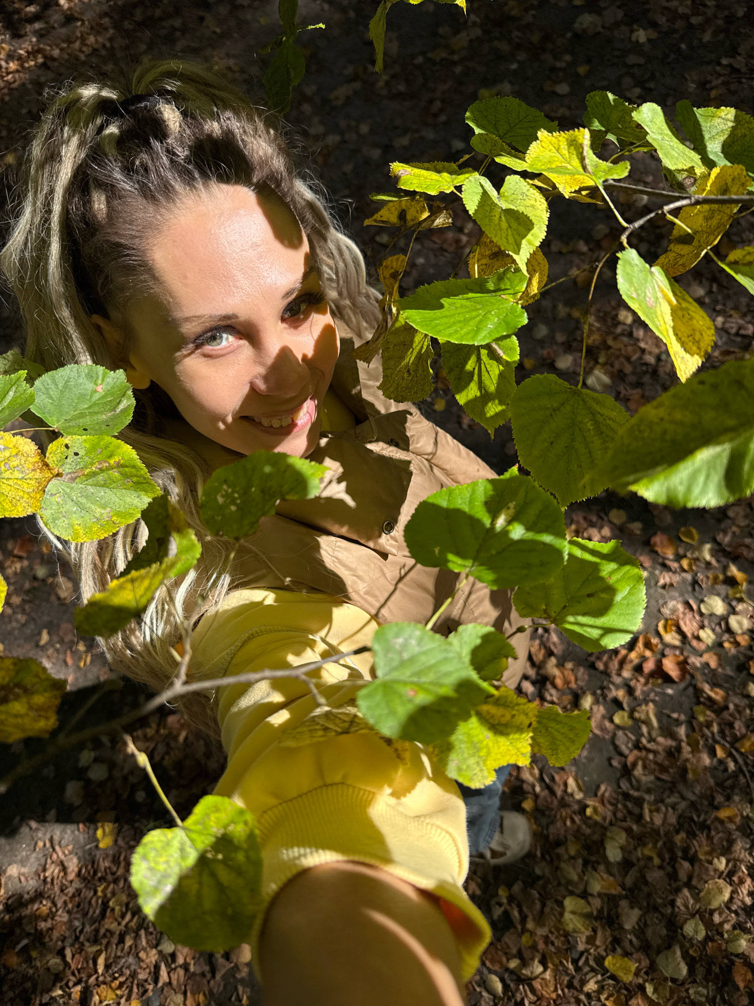 An overhead, close-up shot shows a young woman standing in a forest in Rusanovo, Russia, on a sunny day. She has blonde hair tied up in a ponytail and is wearing a tan jacket over a yellow hoodie. The woman is looking at the camera and smiling, with a branch of green and yellow leaves partially obscuring her face. The forest floor is covered in fallen leaves.