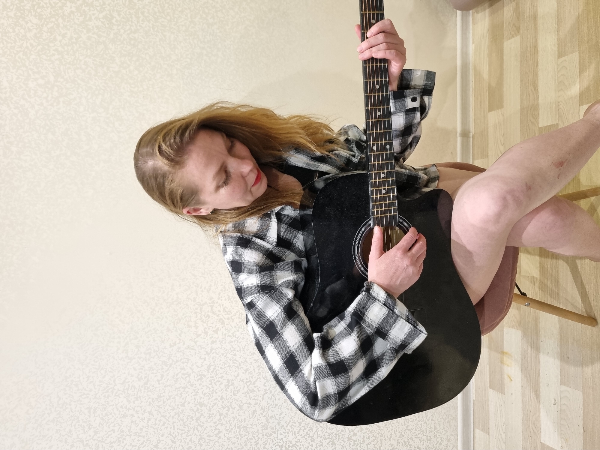 A woman with reddish-blonde hair, wearing a black and white plaid shirt, sits on a chair and holds a black acoustic guitar. She looks down towards the guitar. Her legs are crossed, and a chair leg is visible beneath them. The background consists of a textured wall and a wooden floor. The location is Chaikovsky, Russia.