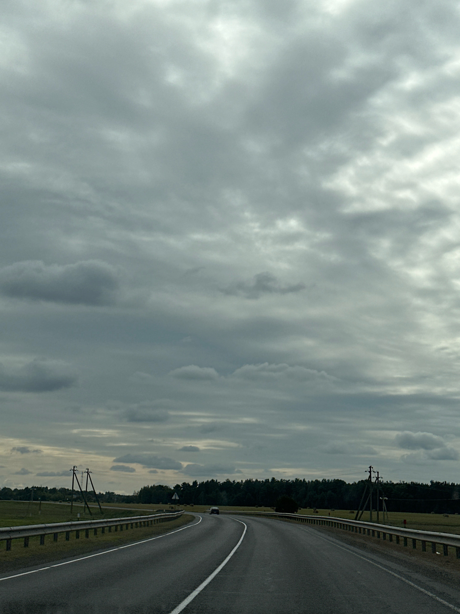 The image captures a view from a vehicle traveling on a two-lane asphalt road in Сычкаўскі сельскі Савет, Belarus. The road curves to the right, bordered by a metal guardrail on the left and a grassy shoulder with hay bales and trees on the right. A single dark-colored car is visible in the distance on the right side of the road. Utility poles with wires are present on both sides of the road. The sky is overcast with heavy, gray clouds, suggesting a cloudy or potentially rainy day. The surrounding landscape consists of fields and a dense treeline in the background.