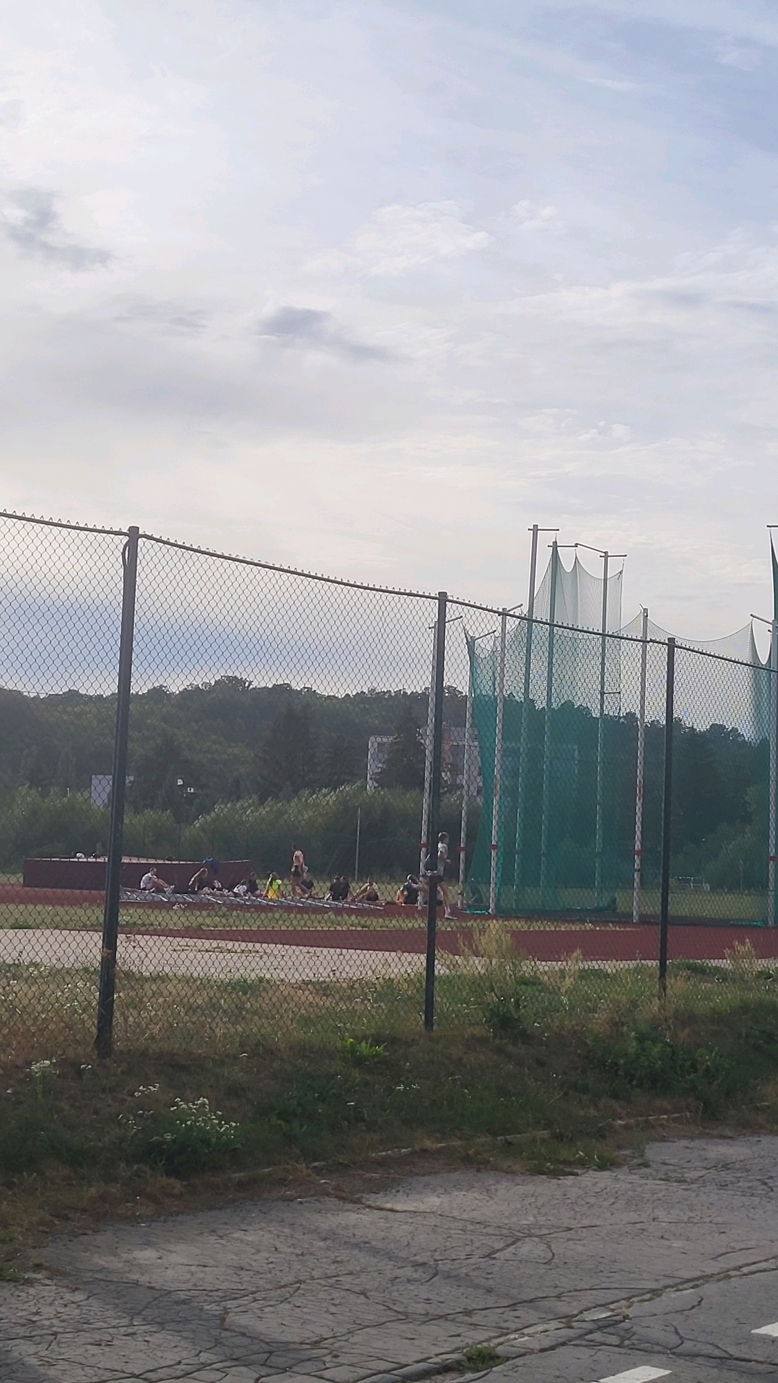 In the city of Prešov, Slovakia, an outdoor athletic facility is depicted, likely a track and field area. A chain-link fence runs horizontally across the foreground, partially obscuring the scene beyond. Behind the fence, a group of individuals, appearing to be young people, are gathered on a red track surface. Many are seated or in a lying-down position, possibly engaged in practice or rest. Some are wearing athletic attire. In the background, a tall green netting structure, characteristic of a throwing area for sports like hammer or discus, stands prominently. Beyond the netting and track, a dense treeline forms the backdrop, with a building or structure visible amongst the trees on a hill. The sky above is overcast with diffused light.