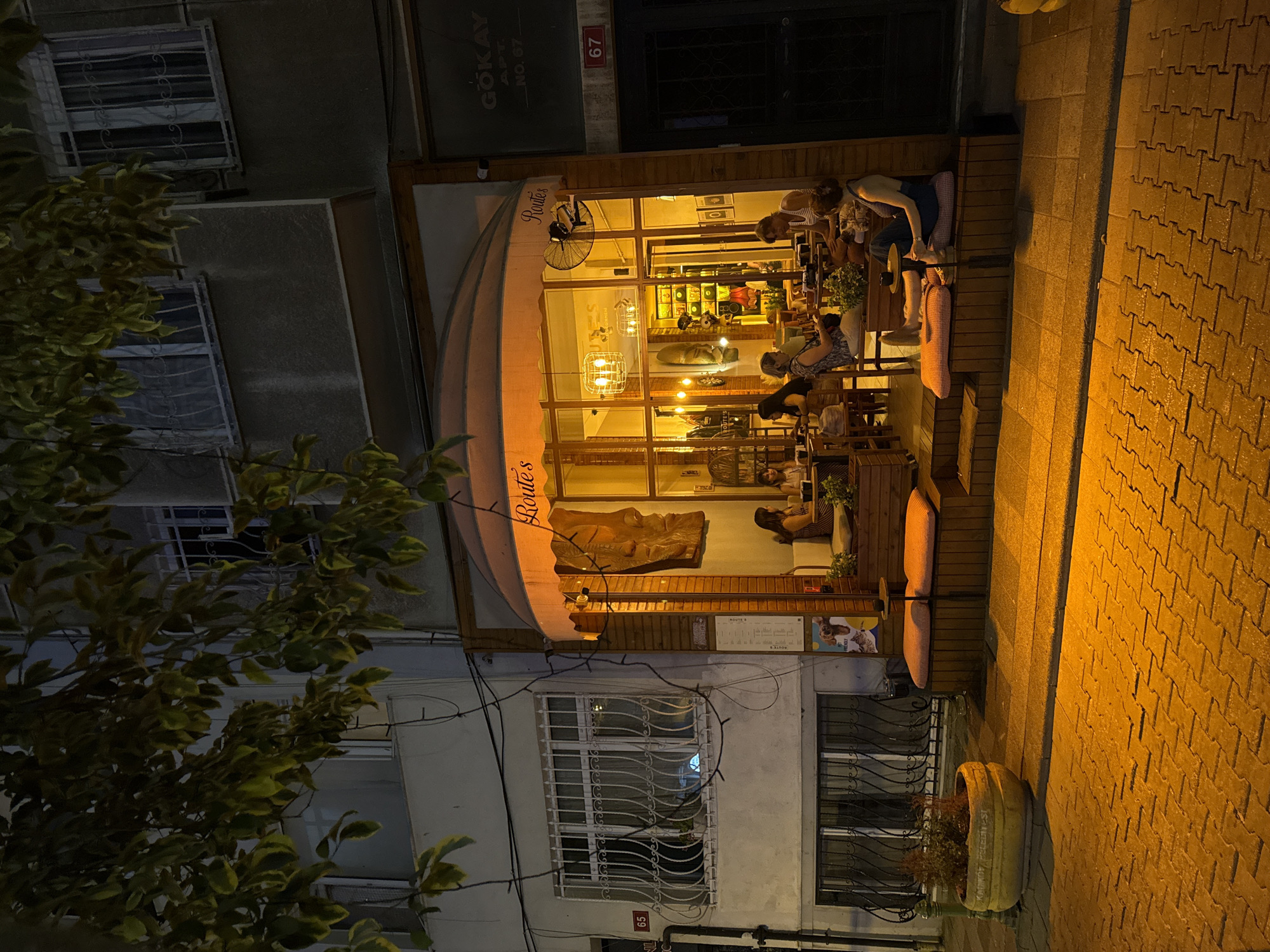 The image captures an outdoor seating area of a cafe at night on a street in Kadıköy, Turkey. The cafe, named "Route's," has large glass windows illuminated from within, revealing tables, chairs, and patrons. Outside, several people are seated at tables and on a raised wooden platform. One woman is seated on a cushion on the platform, leaning back with her legs extended and looking towards the right. Another woman is seated at a table to the left, facing away from the camera. A man and two women are seated at a table inside the cafe.
The cafe is situated in front of a multi-story building. The building's facade features barred windows on the upper floors and a prominent number "65" on the left side. The entrance to an adjacent building is visible on the right, marked with "GÖKAY APT. NO: 67" and the number "67" in a red frame. The street in front of the cafe is paved with textured bricks and is lit by the warm glow of the cafe's lights and streetlights, casting long shadows. A large potted plant with a decorative inscription is placed on the left side of the sidewalk. Overhanging branches of a tree with green and yellowing leaves frame the top of the image. The overall atmosphere is cozy and inviting, suggesting a relaxed evening at the cafe.