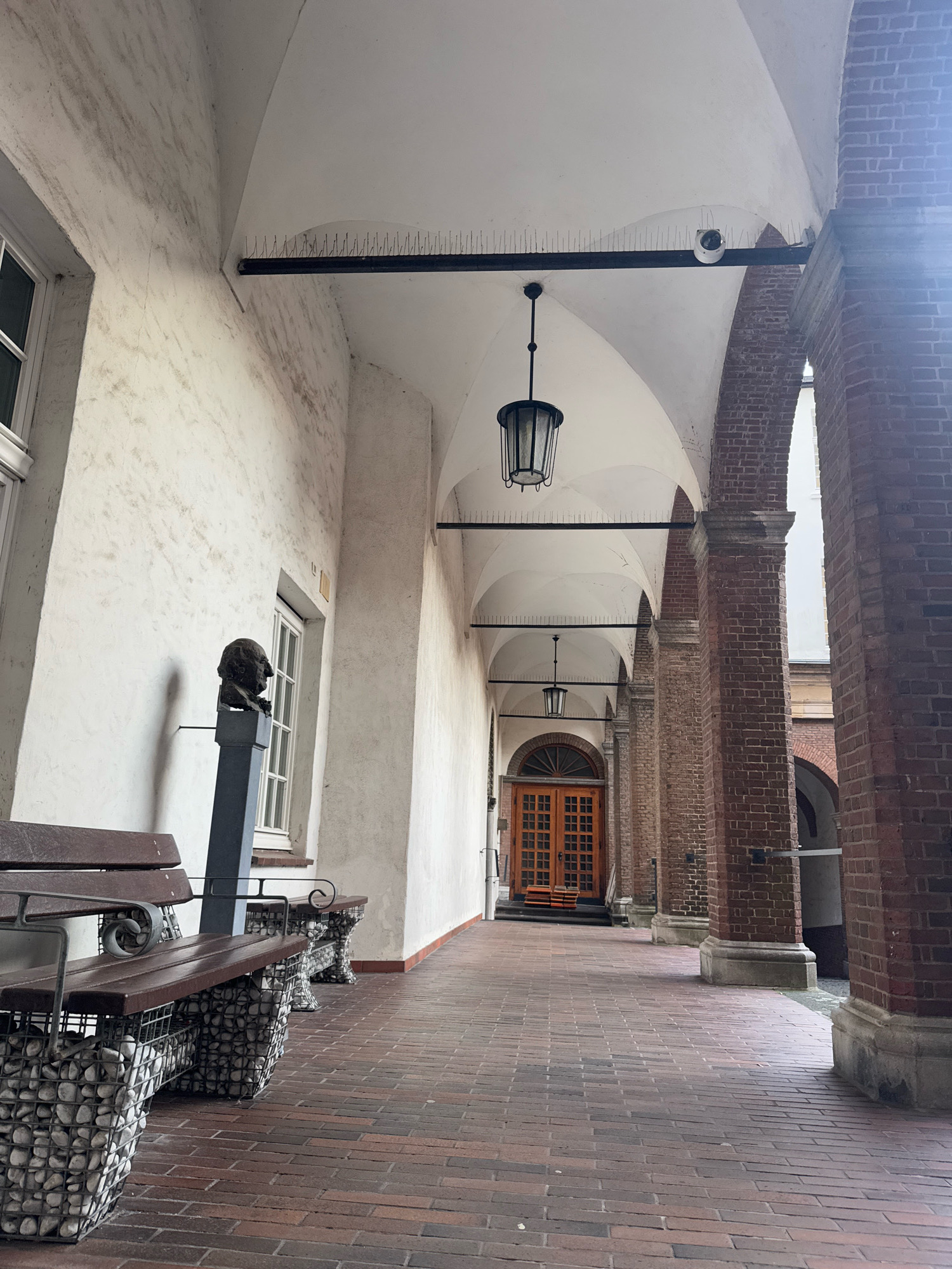 The image depicts an outdoor walkway with an arched ceiling, supported by brick columns. The walkway is paved with red brick. On the left, there is a white wall with a window and a bust of a man on a pedestal. In the foreground, there are two wooden benches with bases made of wire cages filled with stones. Hanging from the ceiling are two lanterns. In the background, a set of wooden double doors is visible. The scene appears to be in a courtyard or a cloister. This location is identified as the inner courtyard of the Museum Kurhaus in Kleve, Germany.