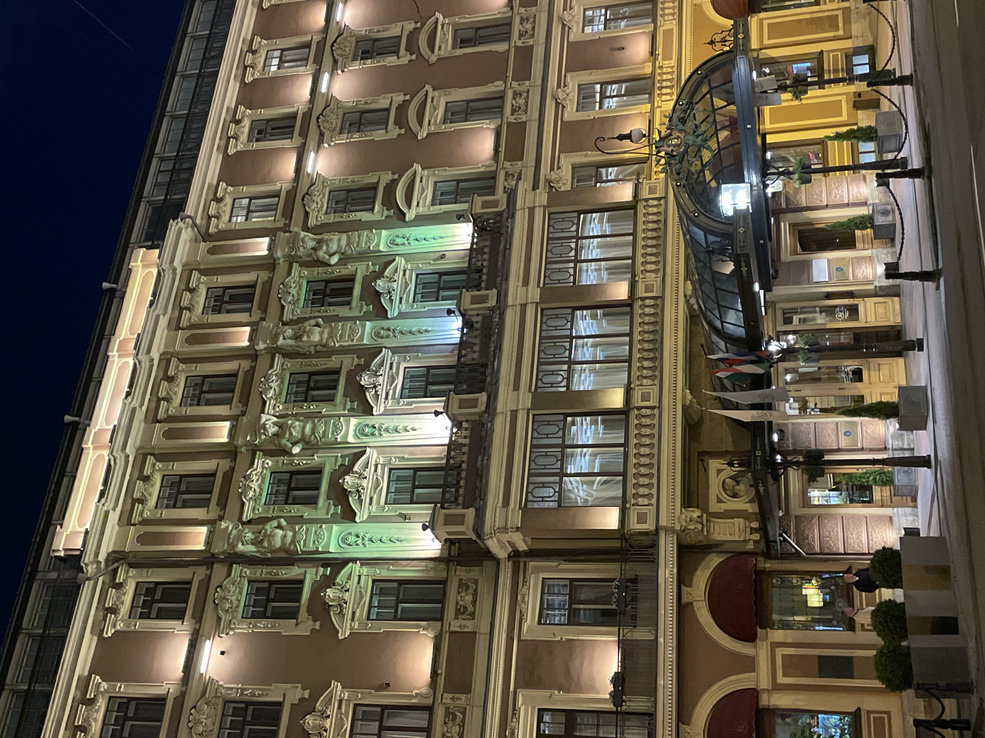 The image shows a tall, ornate building facade in Saint Petersburg, Russia, taken at night. The building is multi-storied, with a mix of architectural styles. The windows are illuminated, and the exterior features decorative carvings and moldings. The lighting scheme emphasizes the building's architectural details. An arched glass-covered entrance is visible on the right, leading into the building. Flags and greenery are placed near the entrance. People are visible in the reflection of a window in the ground floor. The sky is dark blue.