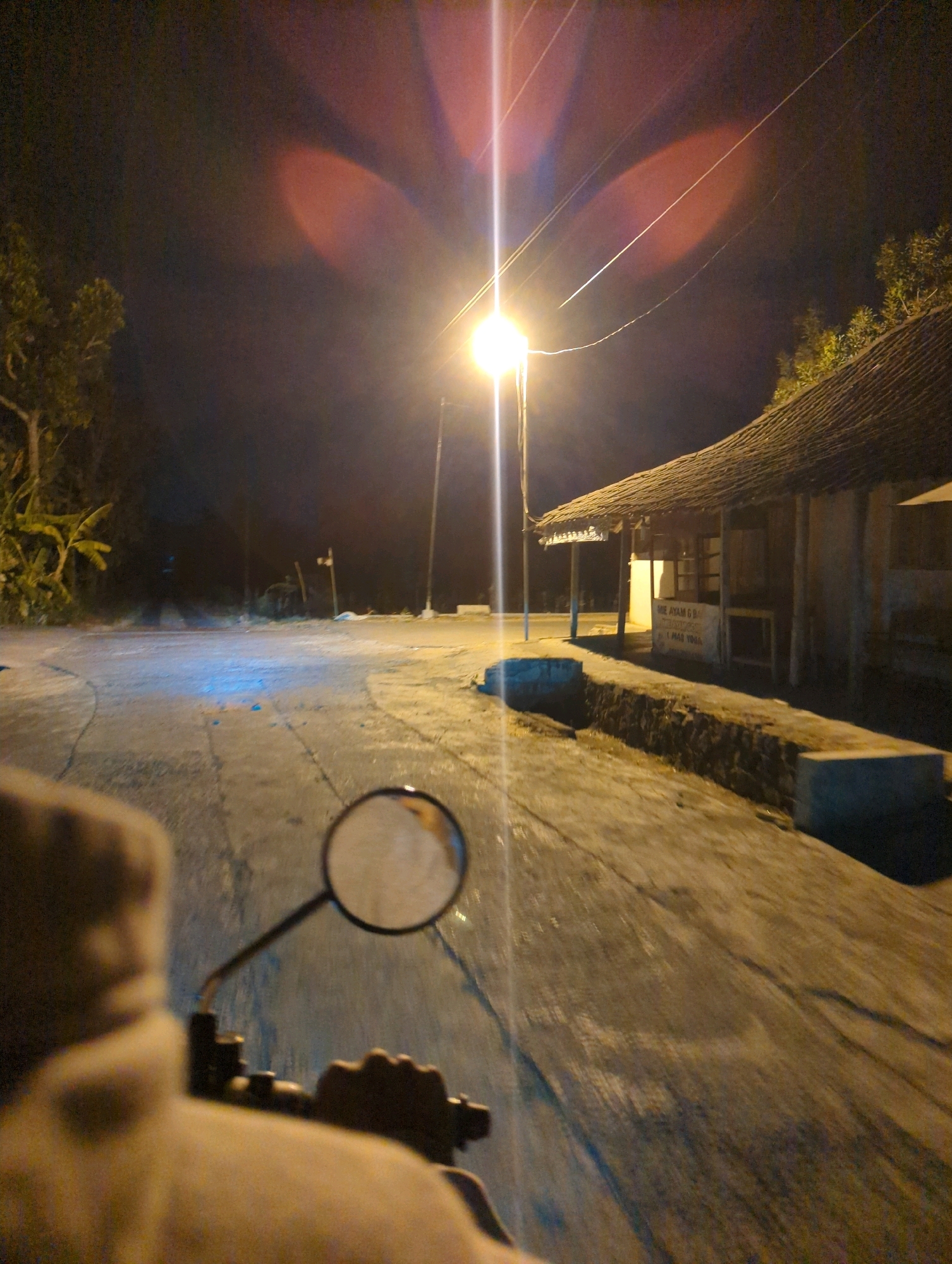 The image shows a nighttime scene on a road, likely in Senggrong, Indonesia, illuminated by a streetlamp. The perspective is from someone riding a motorcycle, with the handlebars and a portion of their clothing visible in the foreground. The road surface appears to be asphalt, with some cracks and variations in texture. To the right, there is a small building with a thatched roof and an open-fronted area that looks like a stall or small shop. Text is visible on the building, partially obscured, but appears to be in Indonesian. Some potted plants or vegetation are visible on the left side of the road. The overall atmosphere is dim, with the streetlamp casting a bright light that creates lens flare effects in the upper portion of the image. The sky is dark, indicating it is nighttime.