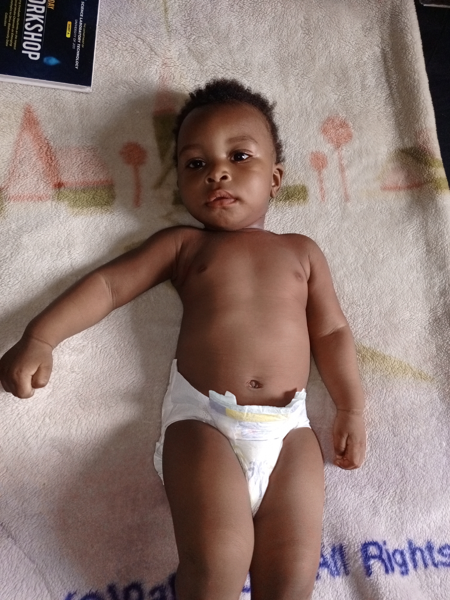 The image shows a baby lying on a patterned blanket wearing a diaper. A book with the title "WORKSHOP" and some text about "SCIENCE LABORATORY TECHNOLOGY UNIVERSITY OF JOS" is visible near the baby's head. The blanket has a light-colored, textured surface with a design featuring flowers or plant shapes in pink and green. "All Rights" is written near the bottom of the image.