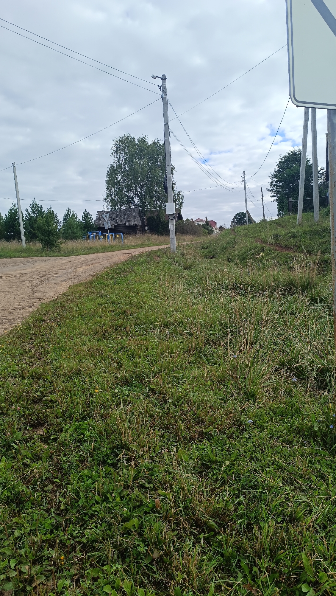 The image shows a rural scene on an overcast day. A dirt road curves to the left, bordered by tall grass on the right. In the background, a dilapidated wooden house with a tin roof is visible, surrounded by trees. Utility poles with overhead wires run across the frame. A white sign is partially visible in the upper right corner. The environment appears to be natural with some signs of human habitation.