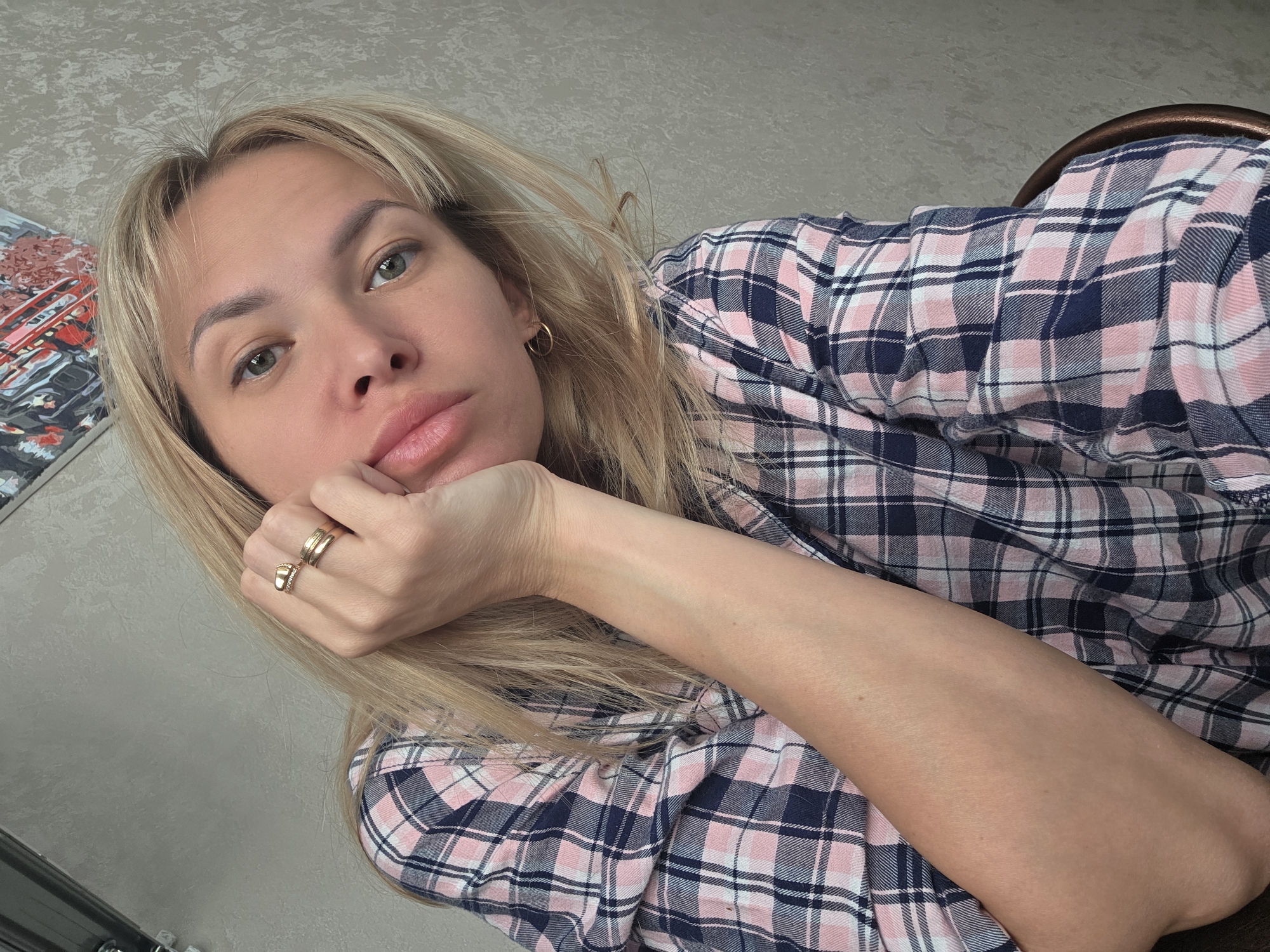 A woman with blonde hair is resting her head on her hand. She is wearing a pink and navy blue plaid shirt and two gold rings on her fingers. The background is a neutral wall, and a piece of artwork is visible in the upper left corner. The location is unknown and there is no information to suggest that it is Chaikovsky, Russia.
