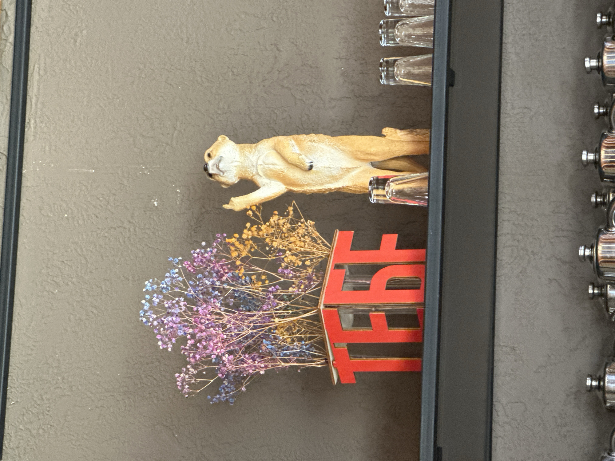 A meerkat figurine stands on a shelf next to a vase of dried flowers. The vase is red and has the letters "TEBE" cut out of it. The shelf is dark grey and is mounted on a textured grey wall. Several small clear glasses are arranged to the right of the meerkat. Below this shelf, another dark grey shelf holds several metal teapots. This scene is located in Krasnaya Polyana, Russia.