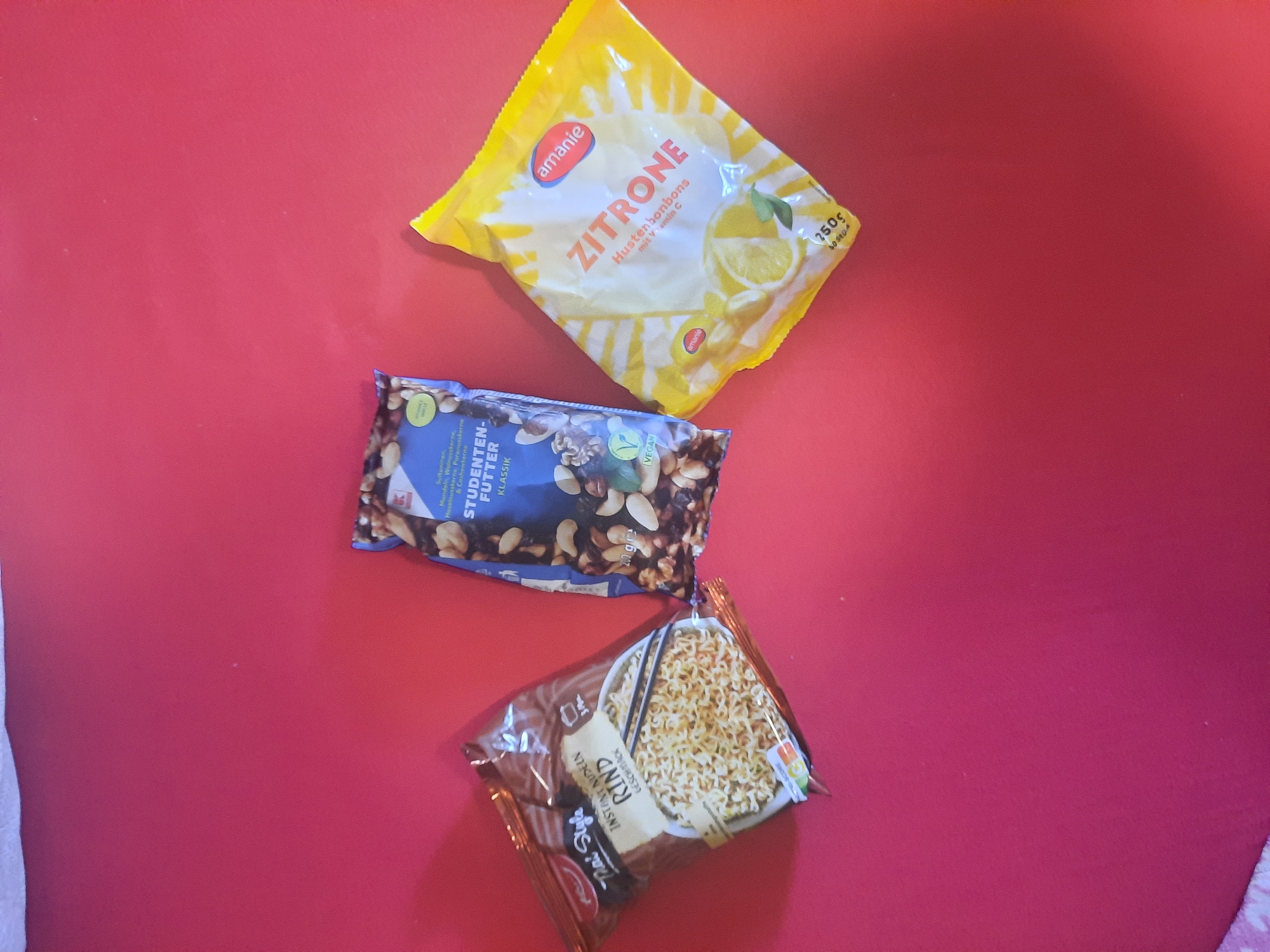 Three food packages are laid out on a red surface. On the left is a package of "Thai Style Instant Nudeln Rind Geschmack." In the center is a package of "Studenten-Futter Klassik" which appears to be a mix of nuts and dried fruit. On the right is a yellow package of "Amanie Zitrone Hustenbonbons mit Vitamin C." The packages are the primary focus of the image, and the red surface provides a simple, uncluttered background.