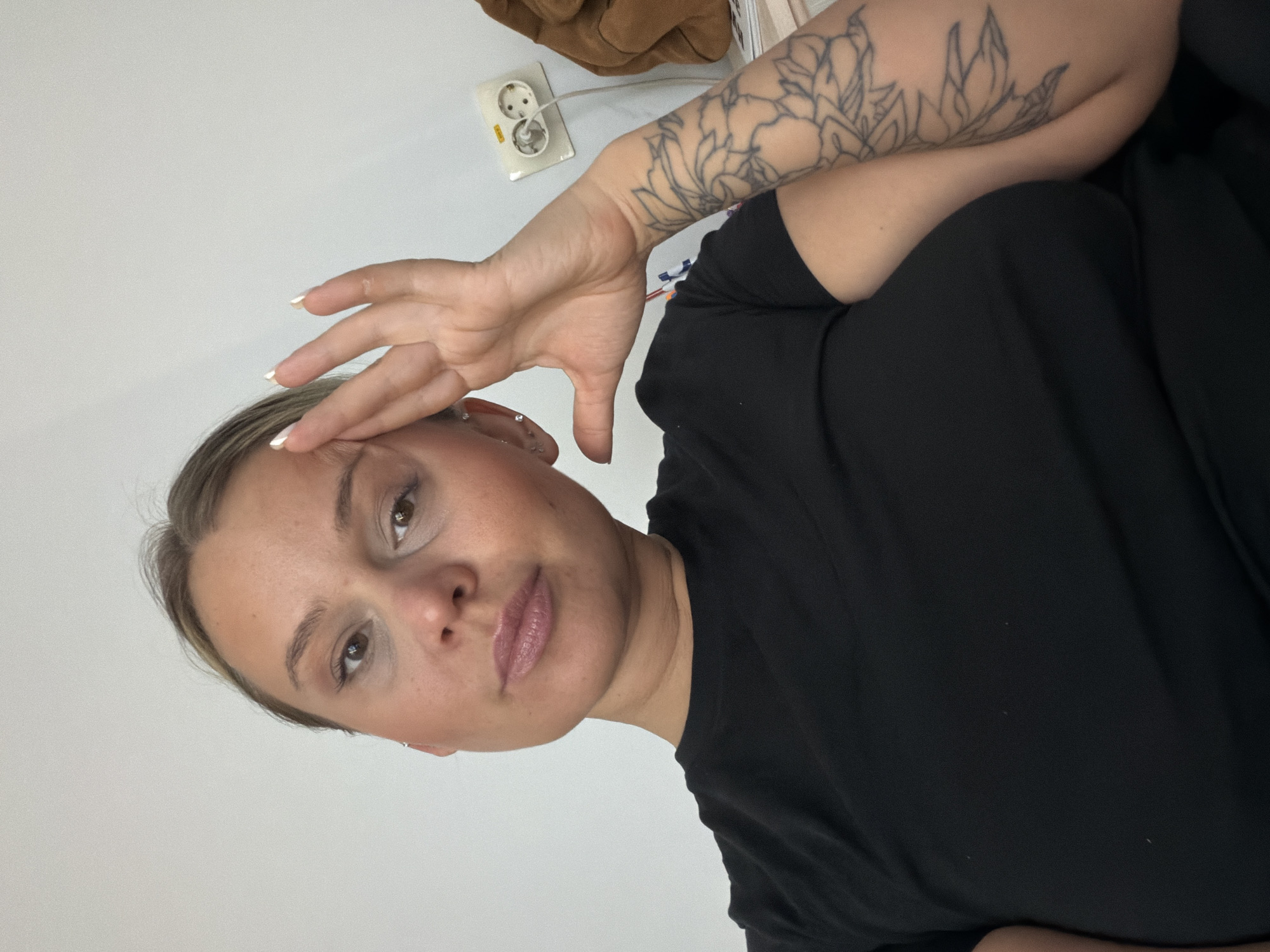 A young woman with light skin and blonde hair, wearing a black shirt, is positioned in the foreground of the image, which is rotated counter-clockwise. Her left arm, adorned with a floral tattoo, is raised, with her hand touching her forehead. She has light brown eyes and neutral-toned lips. In the background, a white wall is visible. A European-style electrical outlet is located on the wall. There is also a brown object and some unidentified white rectangular objects. The image lacks specific indicators linking it to Sarapul, Russia, making a precise location within the city impossible.