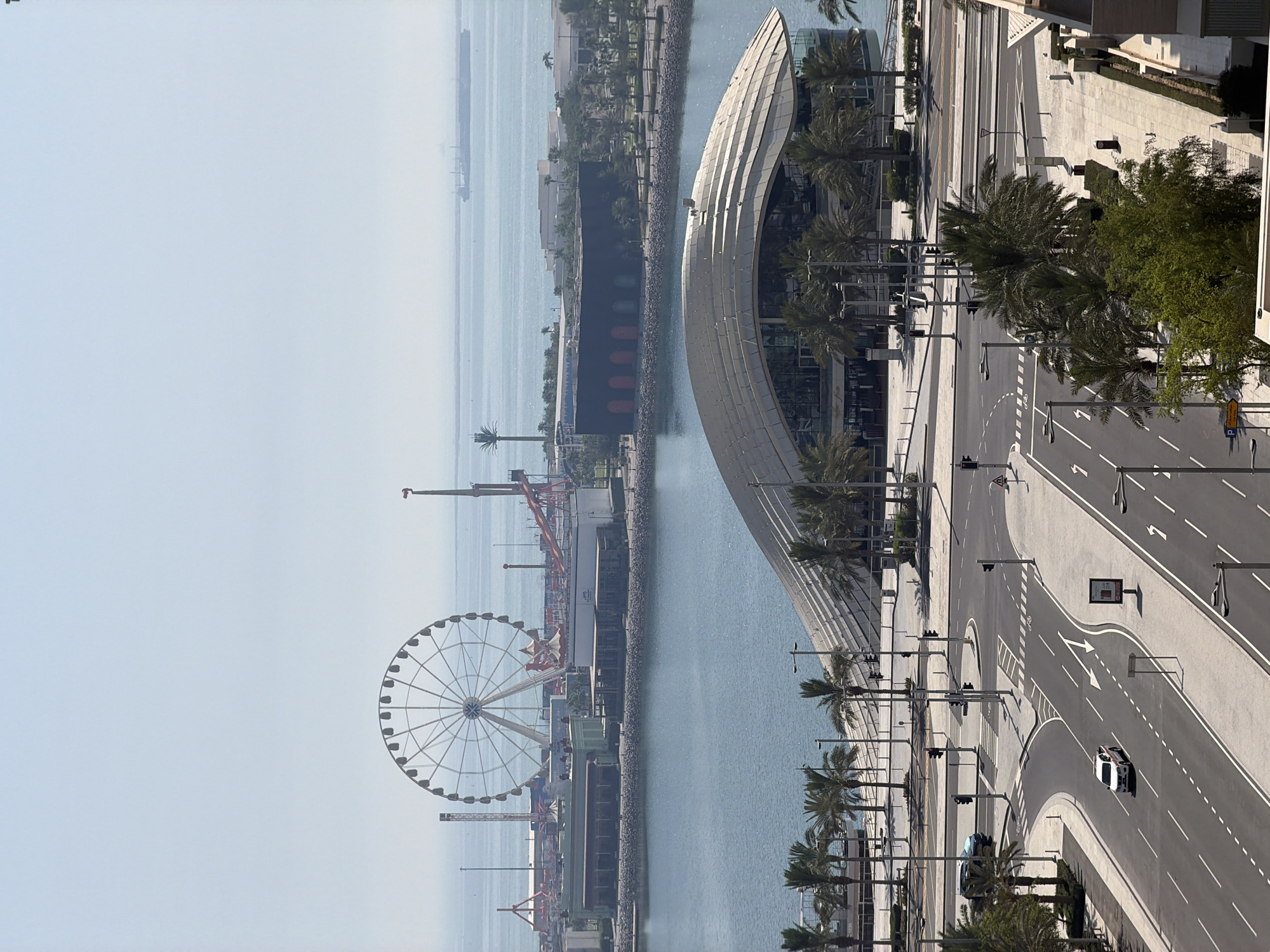 An aerial view captures a coastal city scene under a hazy sky. In the foreground, a multi-lane road with clear lane markings curves through a modern urban landscape. Several cars are visible, including a white car driving on the right lane and a dark SUV parked on the left. Pedestrian crosswalks and traffic lights are strategically placed at intersections. Palm trees line the sidewalks, adding a tropical touch to the environment.
Further in the background, a large body of water separates the foreground from a distinct waterfront area. On the waterfront, a uniquely designed building with a curved, metallic roof stands prominently. Beyond this structure, an amusement park with a towering Ferris wheel and various rides is visible. The scene extends to the horizon, where the calm sea meets the sky, with a large cargo ship visible in the distance. The overall atmosphere is one of a bright, sunny day in a bustling, well-developed urban coastal setting.
