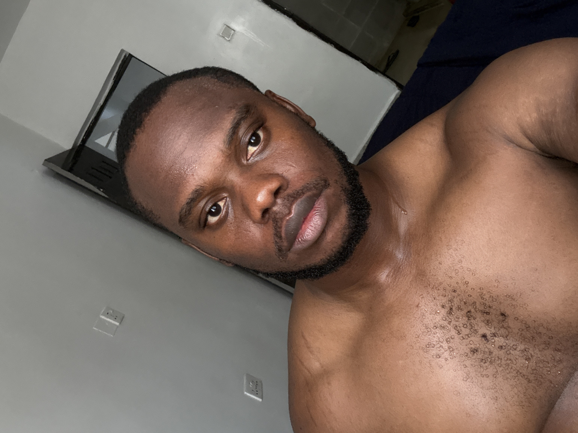 A close-up, angled shot of a shirtless Black man with a trimmed beard. He appears sweaty or glistening with moisture. The background includes a light-grey wall with electrical outlets, a portion of a black electronic device (possibly a laptop), and a dark blue surface (likely bedding). The image does not provide any information regarding the city of Iddo Tudunwada, Nigeria.
