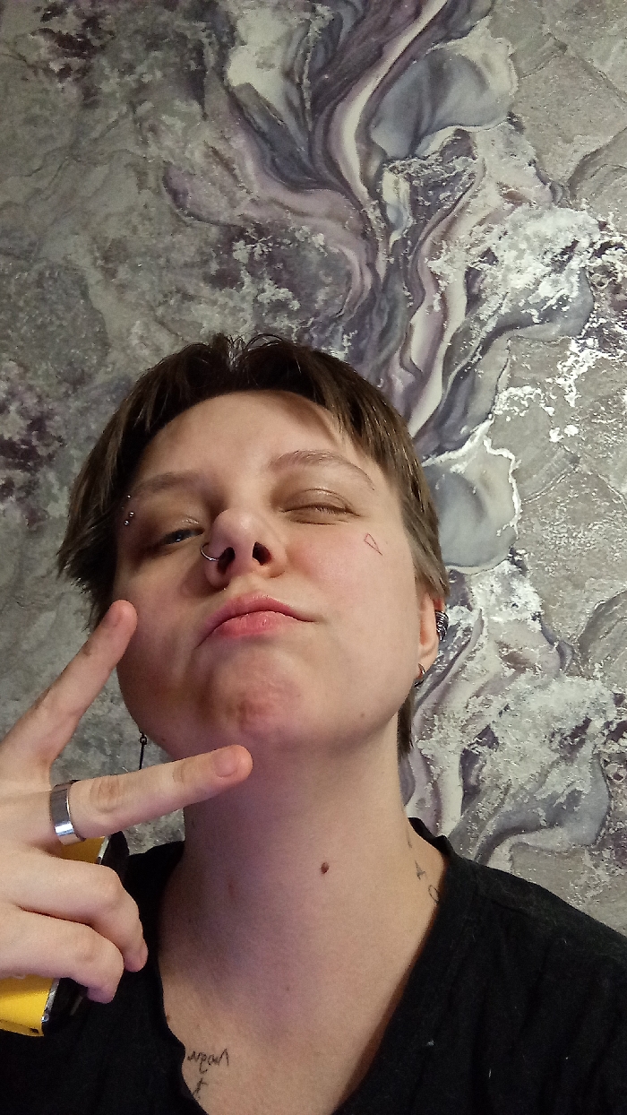 The image captures a close-up portrait of a single individual in an indoor setting in Saratov, Russia. The subject has short, dark brown hair and fair skin. Their right eye (viewer's left) is nearly closed, appearing as a wink or squint, while the left eye (viewer's right) is partially open, revealing a blue iris. Facial piercings include a silver septum ring and three silver studs forming a curved bar on the left eyebrow (viewer's right). A small, inverted triangle outline tattoo is present on the right cheek (viewer's left). The individual's lips are closed and slightly puckered. A small mole is visible on the front of the neck. The subject is wearing a dark-colored, likely black, top. A small, dark tattoo resembling text is visible on the lower left neck/upper chest area (viewer's right). The left hand (viewer's right) is raised, displaying a wide silver-colored ring on the ring finger, with the index and middle fingers extended in a "V" shape. A yellow object, possibly a device, is partially visible, held in the same hand. The background features a wall covered with wallpaper exhibiting an abstract, marbled pattern in shades of grey, white, silver, and subtle purple.
