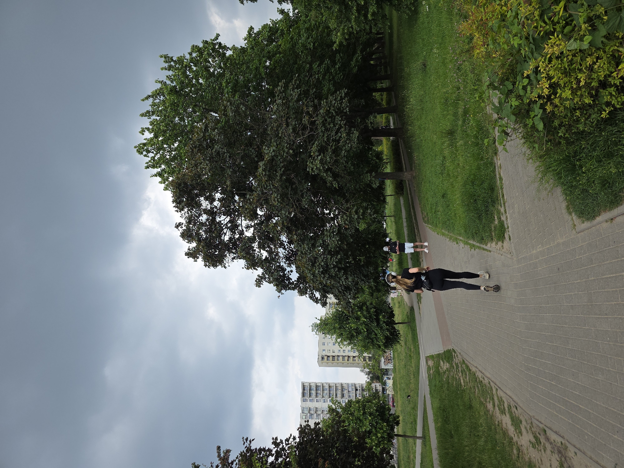 The image depicts a park in an urban setting. Two people, a woman in black athletic wear and a man in shorts and a t-shirt, are walking on a paved path. Large trees line the path, providing shade. In the background, two tall apartment buildings are visible. The sky is overcast with dark clouds.