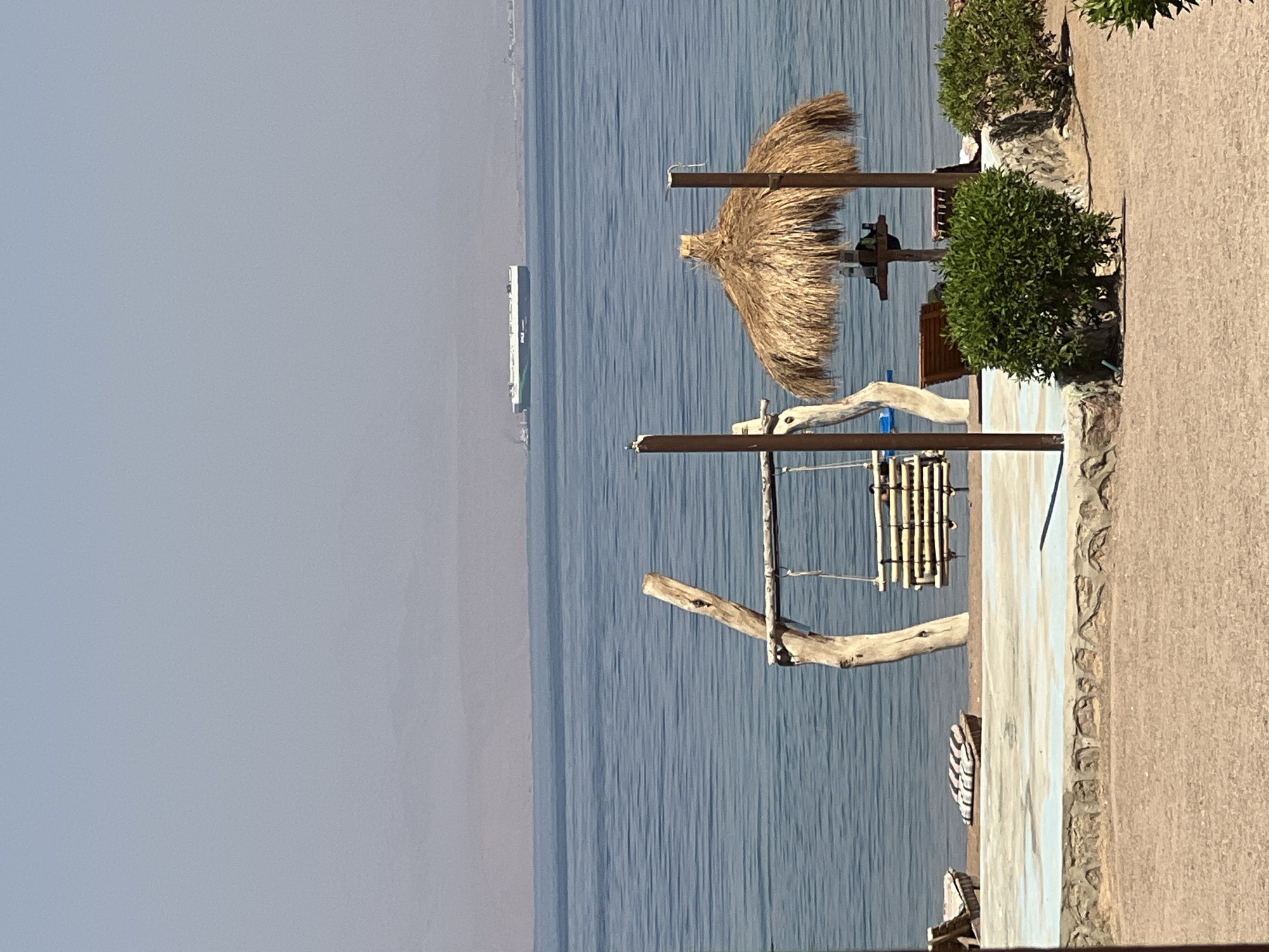 The image captures a serene beach scene in Nuweiba, Egypt, on a clear, sunny day. The tranquil blue waters of the Red Sea stretch towards a hazy horizon, where distant mountains are faintly visible. A large, white cargo ship with green markings is positioned in the middle ground, sailing on the water.
In the foreground, a rustic wooden swing set with a bamboo seat is situated on a sandy shore, adjacent to a thatched umbrella providing shade. Two wooden posts support the swing, and further to the right, the umbrella is supported by a single wooden pole, with a small table underneath. In front of the umbrella, two wooden lounge chairs are visible, and a lush green bush grows to the right of the umbrella. The beach area is bordered by a low, concrete wall with a blue-tinted coping, and beyond that, more sand. The overall impression is one of a peaceful, coastal retreat.