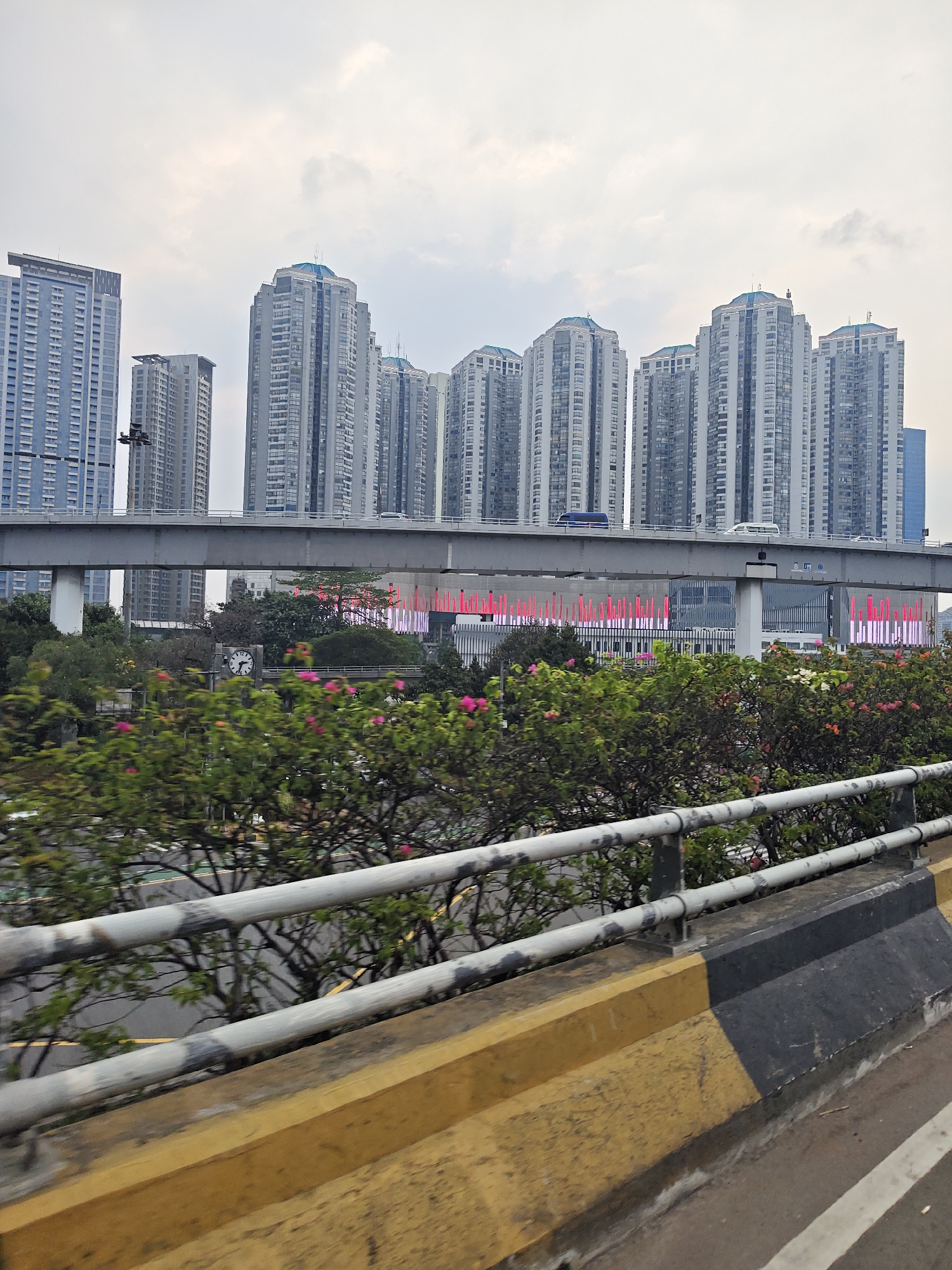 The image captures a cityscape in West Jakarta, Indonesia, characterized by a series of tall, modern apartment buildings. In the foreground, a guardrail with horizontal bars and a concrete barrier painted in yellow and black borders a road. Behind the barrier, lush green foliage and pink flowers are visible. A gray elevated highway or bridge spans across the middle ground, with vehicles, including a blue car and a white van, driving on it. Beneath the highway, a building with vertical red light installations is partially obscured by trees. The sky above is overcast, suggesting a cloudy day. The scene appears to be taken from a moving vehicle.
