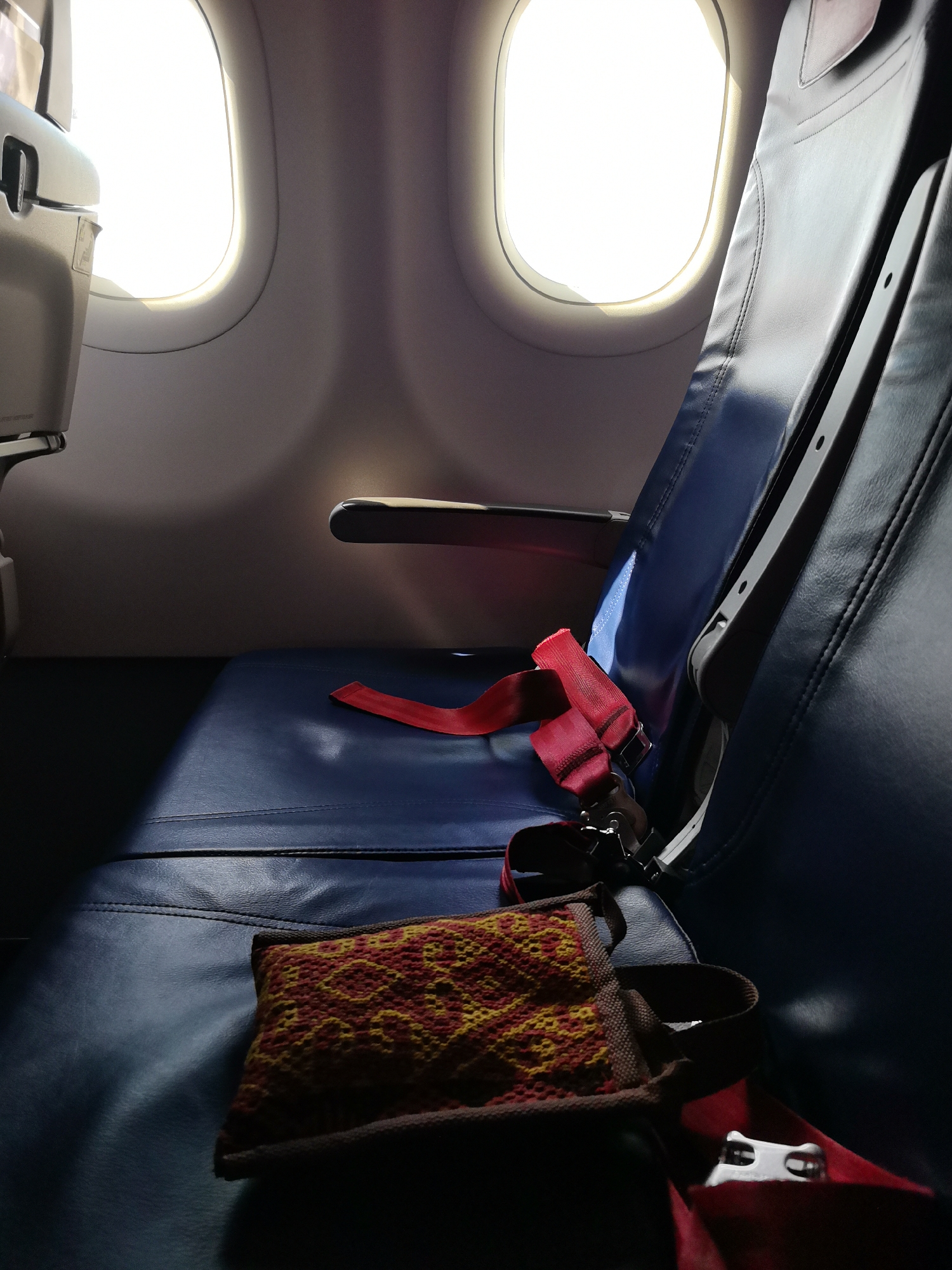 The image shows an interior view of an airplane cabin, focusing on a pair of blue leather airplane seats. A red seatbelt is partially buckled and lies across the seats, with one end draped over the armrest. A small, patterned pouch is placed on the seat in the foreground. Two windows are visible in the background, with bright light streaming through them, casting shadows across the cabin. The image is captured from a low angle, emphasizing the texture and color of the airplane seats and the contrast between the light and shadow. The scene is static and depicts an unoccupied seating area. The location is an airplane cabin, suggesting travel.