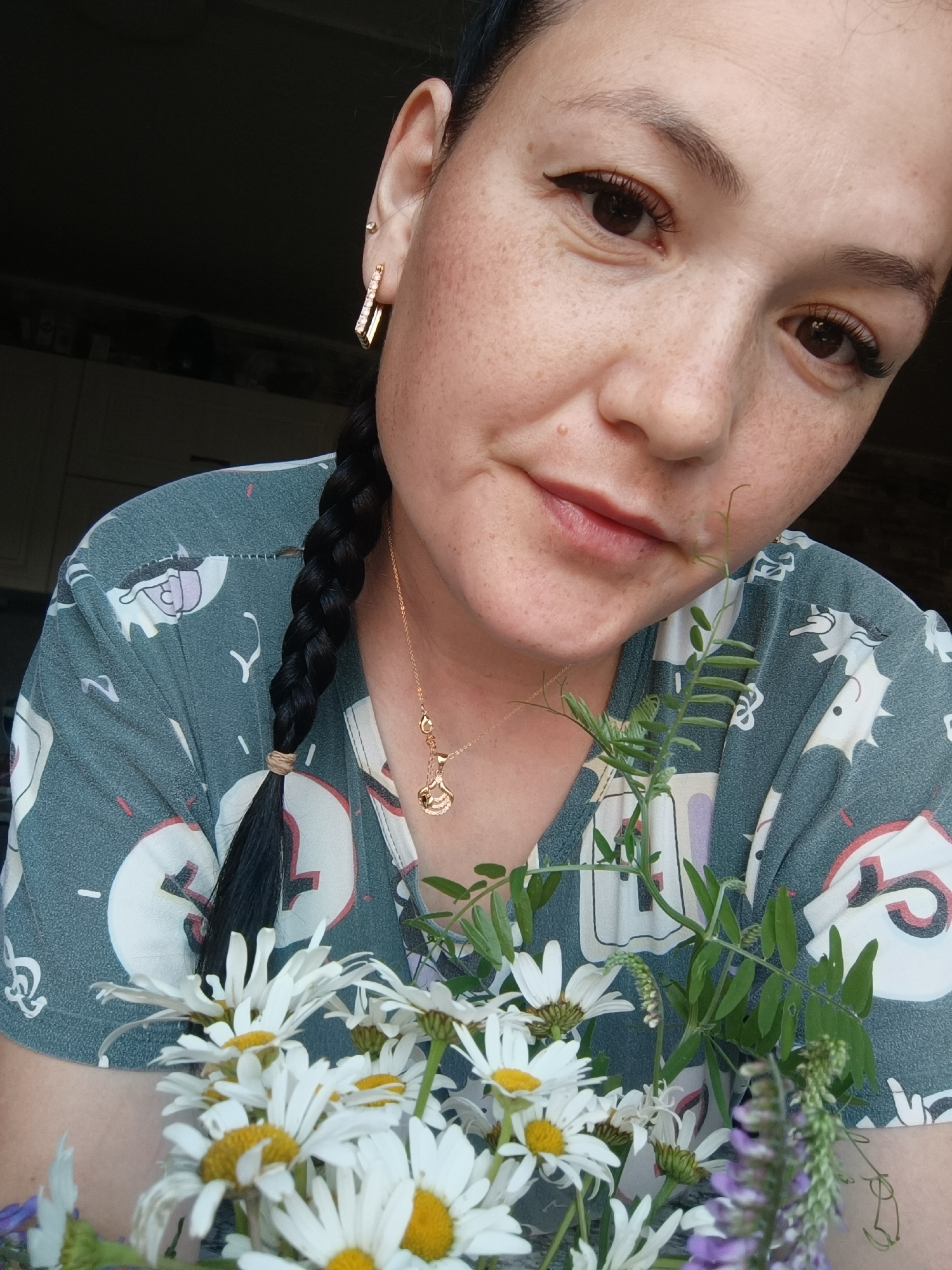 In this close-up, eye-level, portrait-style image, a woman with dark hair styled in a braid is shown from the chest up. She has fair skin with freckles and is wearing eyeliner and mascara. She has a gold necklace with a pendant and gold earrings with white stones. She is wearing a grey t-shirt with a white pattern. She is holding a bouquet of white daisies and other wildflowers. The background is dark and out of focus, suggesting an indoor setting. There is no indication of the city of Karsha, Russia.