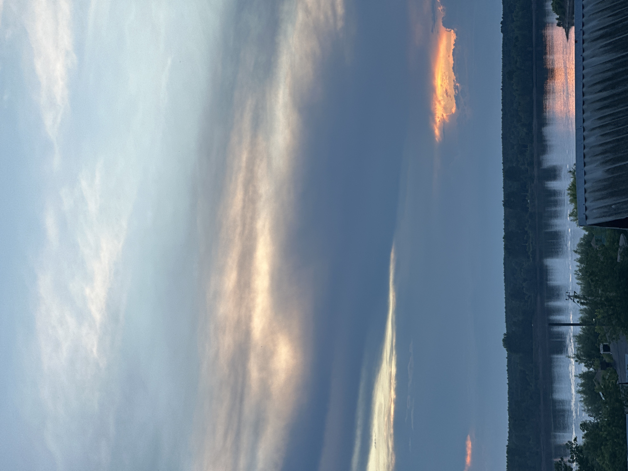 The image captures a wide-angle view of a river and a treeline under a dramatic sky, likely taken from an elevated position within the city of Chaikovsky, Russia. The sky dominates the upper portion of the frame, displaying a gradient of colors from a pale blue at the top to darker blues and grays lower down. Streaks of light orange and yellow clouds are visible, suggesting either a sunrise or sunset. The river occupies the middle ground, its surface reflecting the colors of the sky and showing some disturbance, possibly from wind or boat activity. A dense treeline runs along the far bank of the river, appearing as a dark, continuous mass. In the foreground, the roof of a building, possibly a house, is visible, constructed of corrugated material. Hints of trees and some structures suggest a rural or semi-rural setting on the near side of the river. There are no people or animals clearly visible in the frame. The overall impression is one of a natural landscape at dusk or dawn, with human settlement present but not the primary focus.