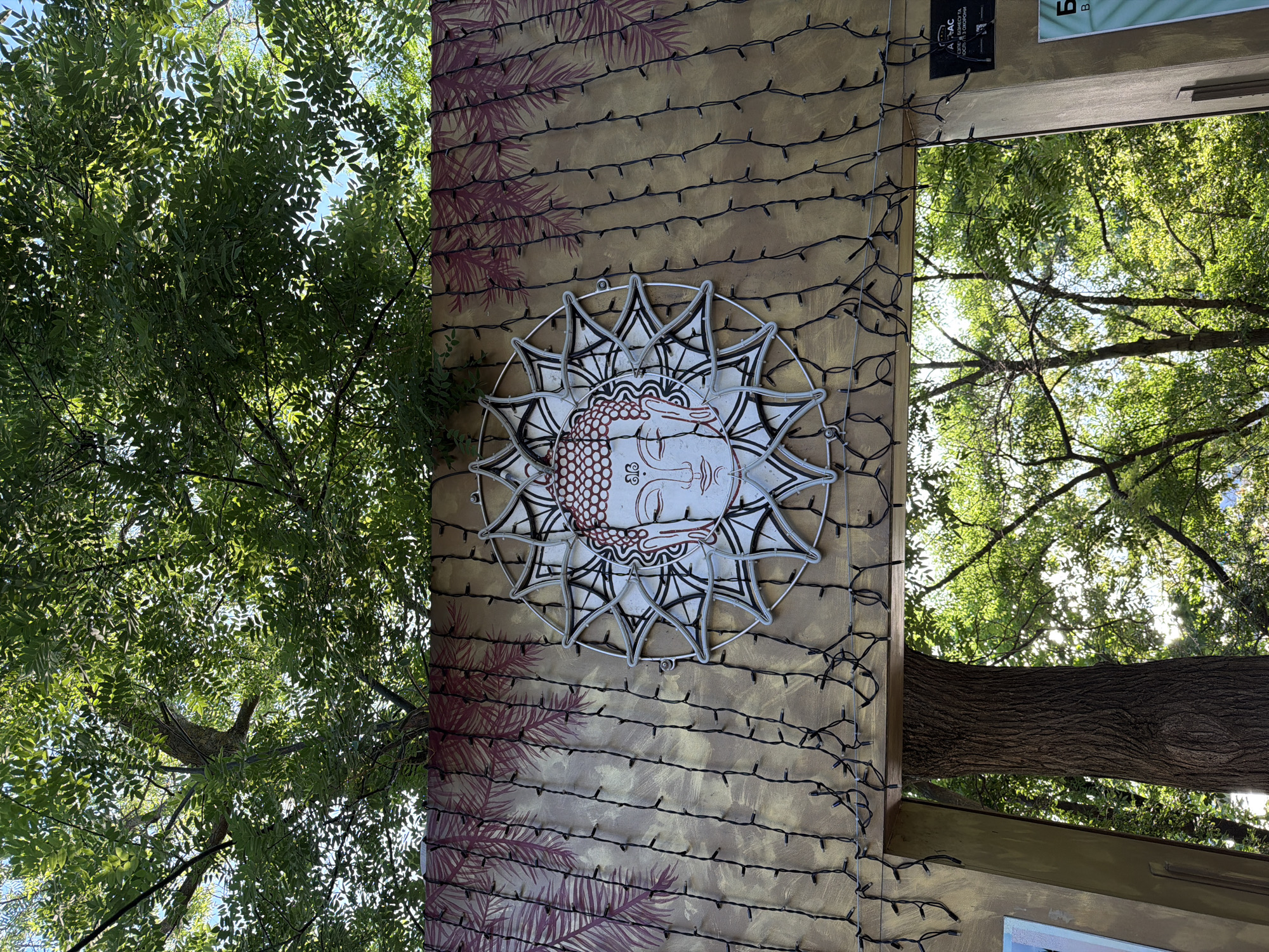 The image depicts an outdoor scene featuring a decorative archway with a Buddha face mandala at its center, adorned with string lights. The background shows lush green trees and a blue sky, suggesting a park or garden setting. There are no people visible in the frame. The archway itself appears to be constructed of wood and is decorated with a backdrop featuring palm leaf motifs. A sign with Cyrillic text and a logo is visible on the right side of the archway. The overall impression is one of a tranquil and decorative outdoor space. The image was taken in Odesa, Ukraine.