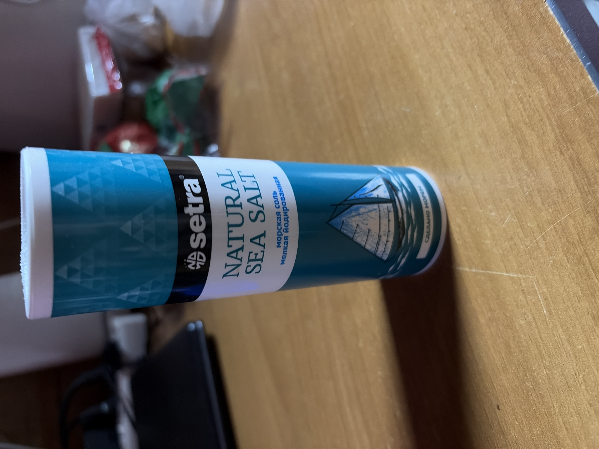 The image shows a can of "Setra" brand natural sea salt lying horizontally on a wooden surface. The can has a blue and white color scheme with a sailboat design. The text on the can is in both English and Russian, indicating it is "Natural Sea Salt" (in English) and "Морская соль мелкая йодированная" (Sea salt fine iodized in Russian). The background is slightly blurred, but suggests a domestic setting. Based on the text on the can, it appears to be produced in Russia, but it is impossible to determine the exact location in the city of Staryi Menzelyabash from the visual information provided.