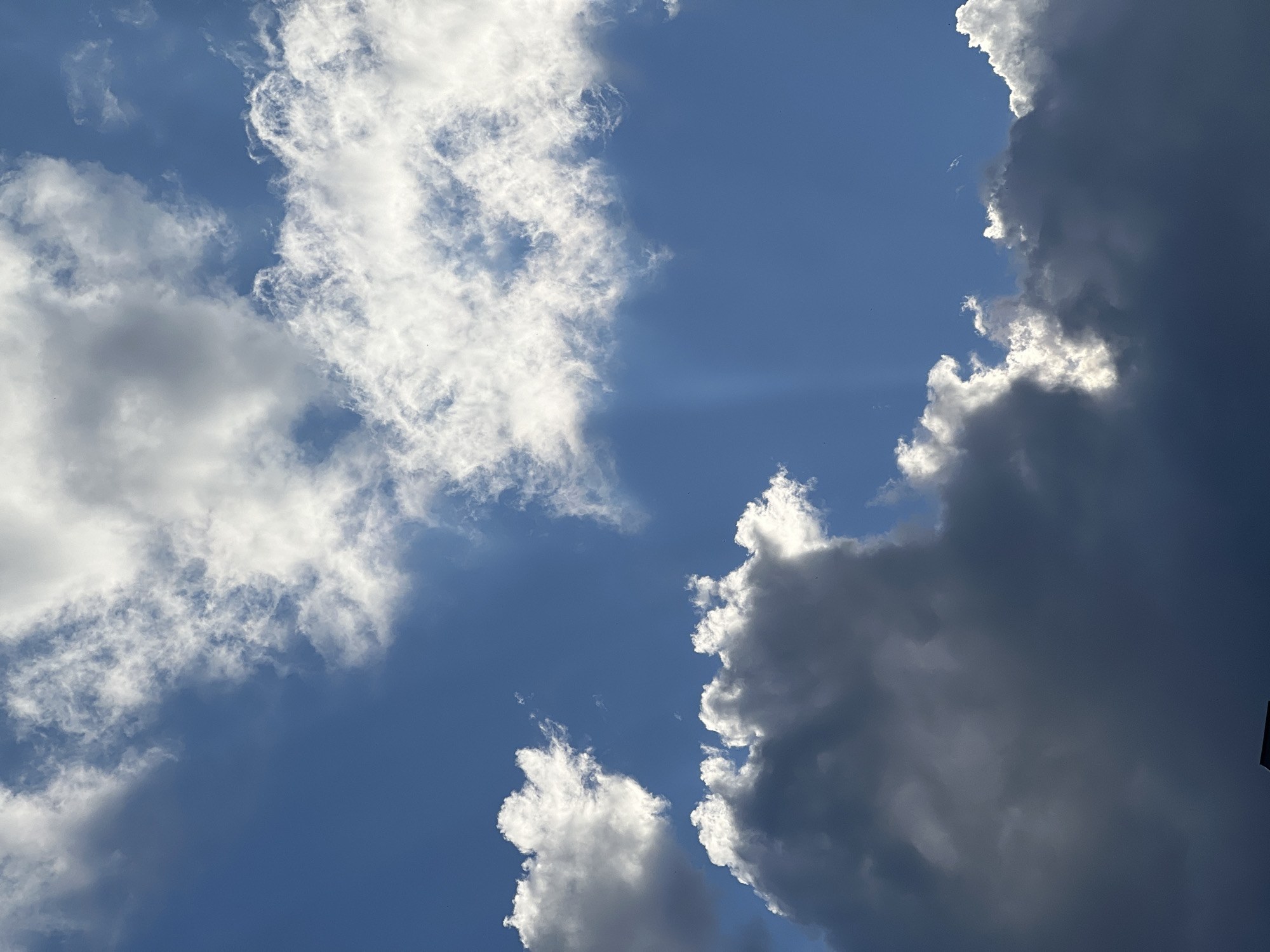 A cloudy blue sky is visible in Yekaterinburg, Russia. The clouds are a mix of white and dark gray, with sunlight peeking through in some areas. A faint beam of light is visible in the upper right portion of the image. In the lower portion of the image, the dark silhouette of a building is visible against the sky. There are no people or animals present in the image. The scene appears to be an outdoor shot, possibly taken during the day.
