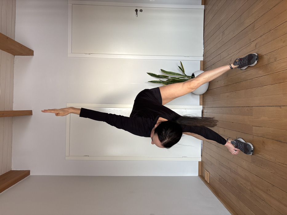 An individual is depicted indoors, performing a yoga or stretching pose. The person, appearing female, has long dark hair tied in a ponytail and is dressed in a black long-sleeved top, black shorts, and dark athletic shoes with grey and white soles. They are bent forward and to the right, with the left arm extended downwards, hand touching the floor near the left foot, and the right arm extended vertically upwards towards the ceiling, fingers outstretched. The head is tilted upwards, looking towards the raised hand. The environment features light brown wooden floorboards, white walls, and a white ceiling with two exposed light brown wooden beams. Two white doors with dark handles are visible in the background. A tall green snake plant in a white, ribbed pot is located behind the individual's right leg. A small, light-colored rectangular object rests on the floor against the left wall.