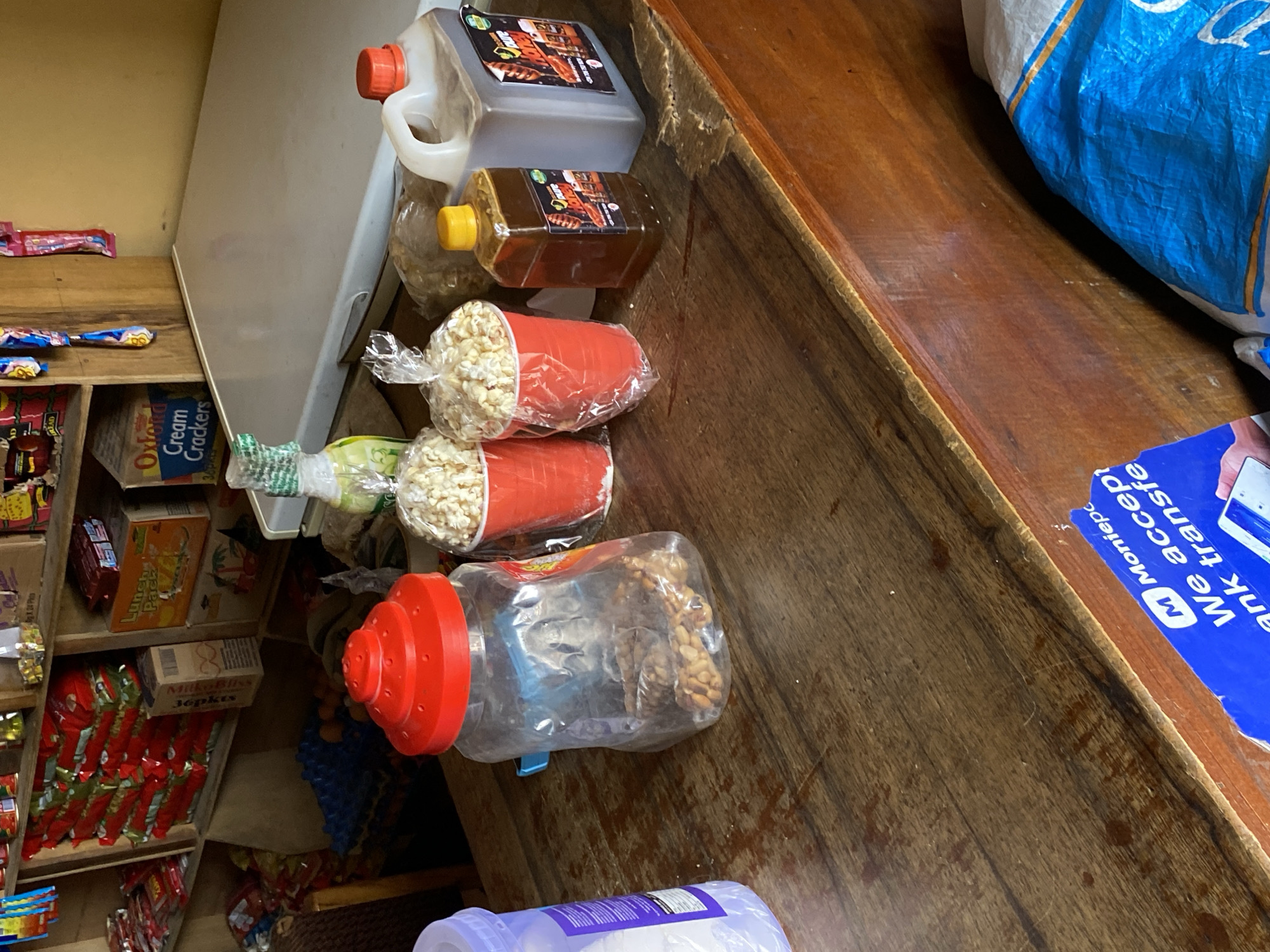 An overhead view captures the interior of a small shop or kiosk, likely located in Bukuru, Nigeria. The scene is dominated by a wooden counter on the right, filled with various packaged goods.
On the counter, two containers of liquid or oil are prominent: a large grey plastic jerry can with an orange cap and a smaller brown rectangular bottle with a yellow cap. Both feature labels with the word "Doure." Below these are two red cups, each filled with popcorn and sealed in clear plastic bags. A clear plastic jar with a red, ridged lid contains brown, fried snacks, possibly peanuts. Part of a blue and white woven bag is visible in the upper right corner. A blue sticker on the counter clearly reads "M Moniepc" (Moniepoint) and "We accept bank transfe," indicating a financial service. A purple plastic bottle is partially visible at the bottom of the frame.
To the left of the counter, a multi-tiered wooden shelving unit is packed with various products. Among them, boxes labeled "Oxford Cream Crackers" and "MaLoBINS 36pkts" (likely Milk Bliss in 36-packet units) are discernible. Numerous other brightly packaged snacks and boxes are arranged on the shelves. A white refrigerator or large appliance is partially visible in the background on the far left. The overall impression is that of a bustling, small-scale retail establishment.