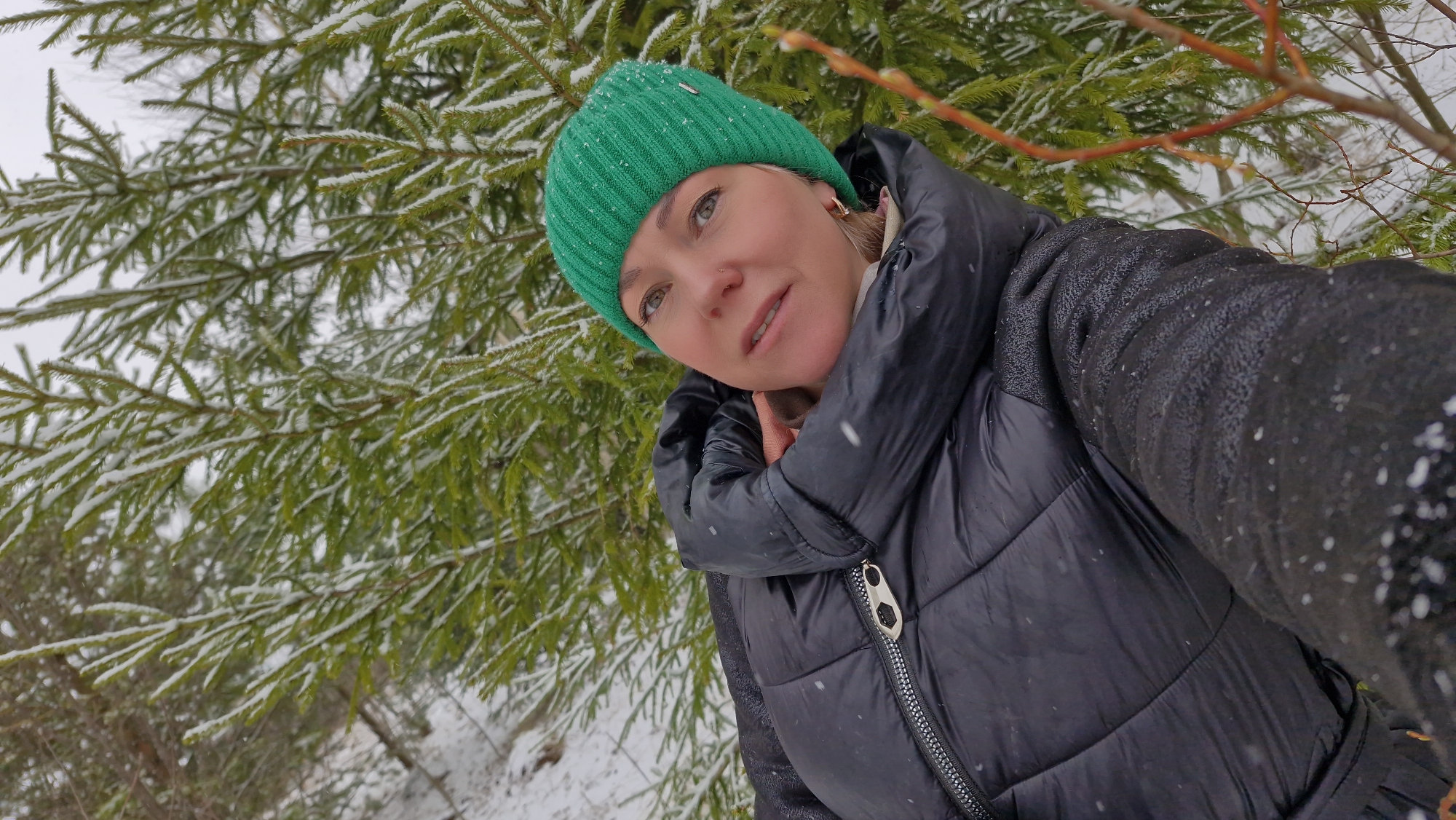 A woman is captured in a close-up, upward-angled shot, posing outdoors in a snowy, wintery setting in Otchor, Russia. She wears a vibrant green knitted beanie and a black quilted puffer jacket with a silver zipper. Her face is turned slightly upwards and to the right, revealing light-colored eyes and a serene expression, with a subtle nose piercing visible. A light-colored collar or scarf is visible around her neck.
The background is dominated by dense green spruce or fir trees, their branches heavily dusted with fresh snow. Some slender reddish-brown branches, possibly from willow or similar deciduous trees, are also visible in the frame. Snowflakes are actively falling and can be seen on her jacket and scattered across the evergreen needles, emphasizing the cold, winter conditions. The ground is covered in snow. The lighting appears soft and diffused, typical of an overcast winter day. The scene evokes a feeling of calm enjoyment amidst the wintry landscape. No visible text is present in the image.