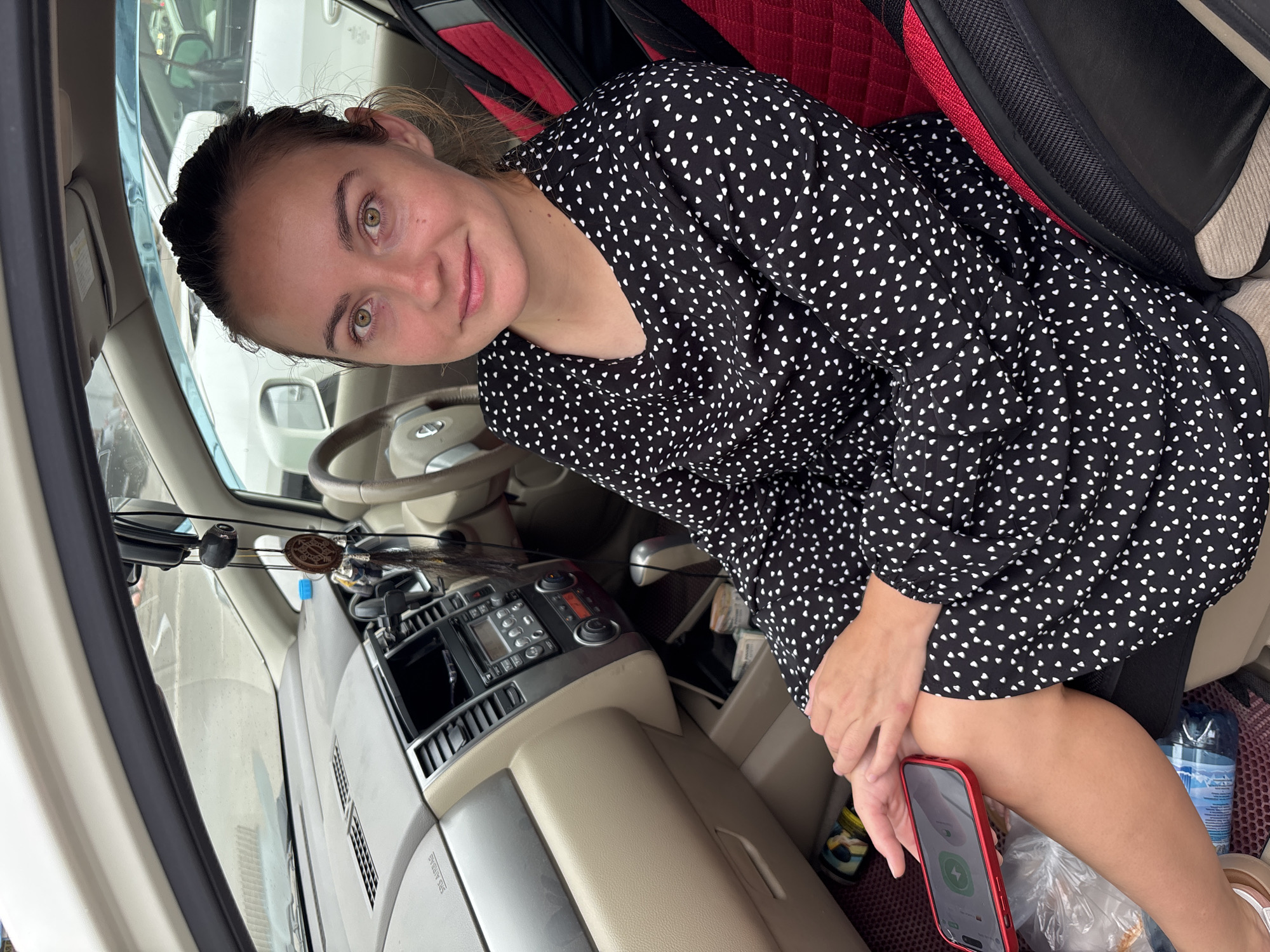 A woman is sitting in the passenger seat of a car, looking at the camera. She is wearing a black dress with white heart patterns and has her hair pulled back. She is holding a red smartphone in her lap. The interior of the car is visible, including the dashboard, steering wheel, and car stereo. The car appears to be parked, possibly in a parking lot, and other vehicles can be seen through the windows. The woman's eyes are a prominent green color. The context of Blagoveshchensk, Russia is not discernible from the visual information provided in the image.