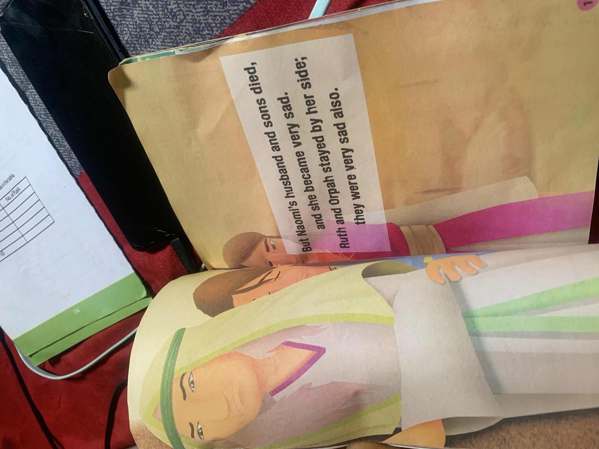 The image shows an open book with illustrations and text. The left page features a colorful illustration of a woman wearing a headscarf and draped clothing, with another figure partially visible behind her. The right page contains text in black font against a light brown background. The text reads: "But Naomi's husband and sons died, and she became very sad. Ruth and Orpah stayed by her side; they were very sad also." A pink stripe runs vertically on the right page, behind the text. The book is placed on a textured surface, possibly a rug or blanket, in shades of grey and blue. In the background, a portion of a green and white notebook or folder is visible, with some handwritten lines and numbers. A white cable runs from the top left of the image towards the notebook. A black smartphone with a cracked screen is also visible behind the book. The lighting suggests an indoor setting. There is no information in the image to suggest a location in Jauro Yinu, Nigeria.