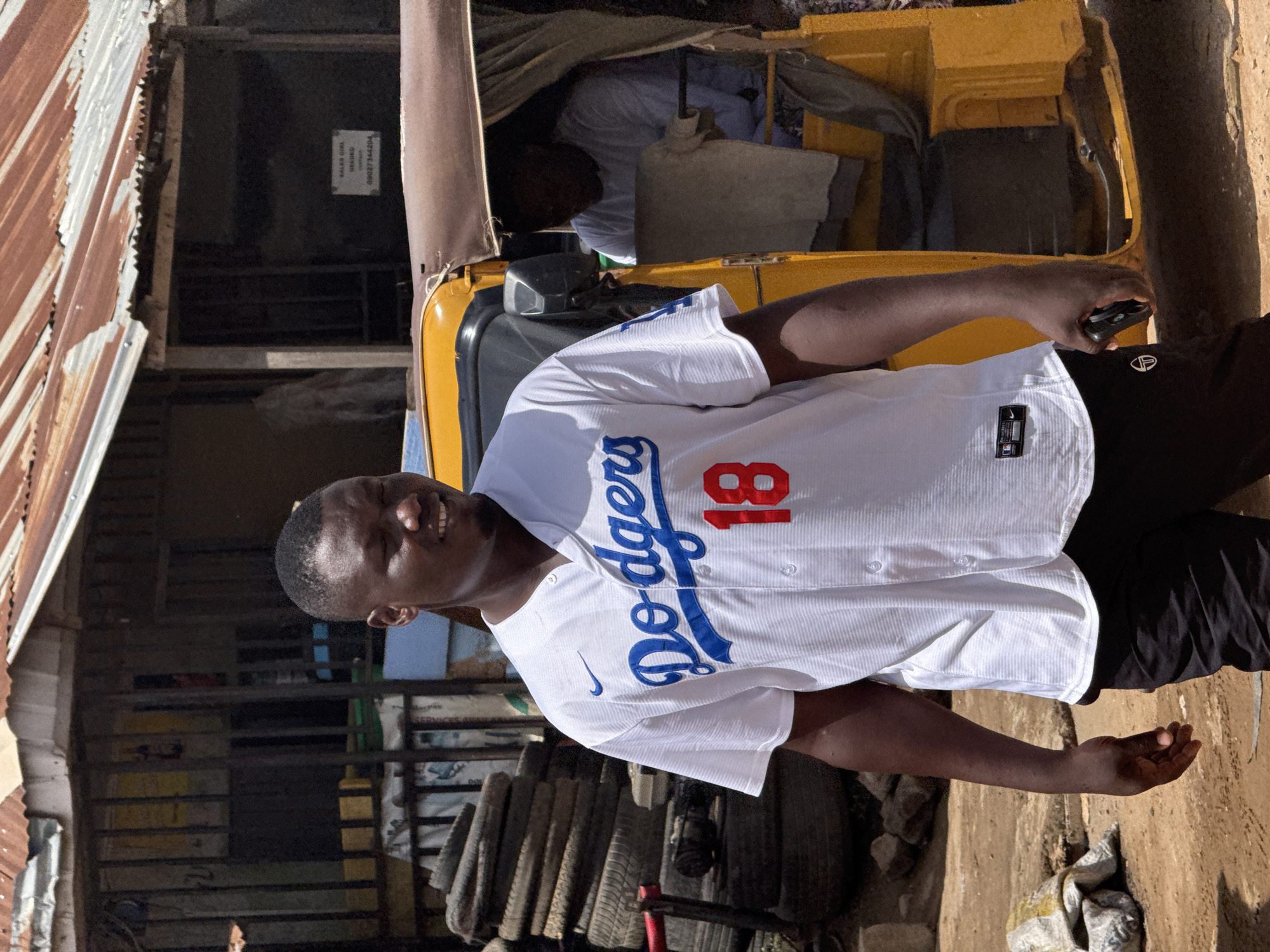 A man wearing a white Dodgers jersey with the number 18 is standing next to a yellow commercial tricycle (Keke Napep) in what appears to be an urban area. The location is likely in Abuja, Nigeria. Behind the man is a building with a corrugated metal roof and a sign with text and a phone number. There are several tires stacked behind him. The ground is paved and there is some debris near the edge.