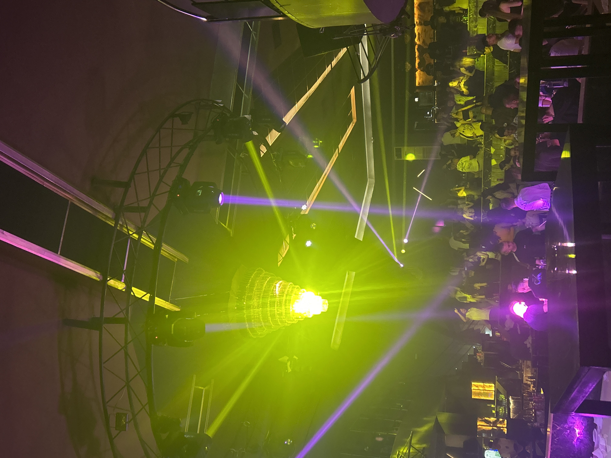 The image captures a lively indoor scene, possibly a nightclub or a bar, located in Zlatograd, Bulgaria. On the right, blurred figures of people are visible, seated at tables or standing at the bar, indicating a social gathering. The left side features a complex lighting setup with a large, circular metal structure holding various spotlights. A chandelier-like lighting fixture emits a bright, central glow surrounded by beams of green and purple light, which illuminate the space. The environment is dimly lit with a focus on the lighting effects, creating a dynamic and festive atmosphere.