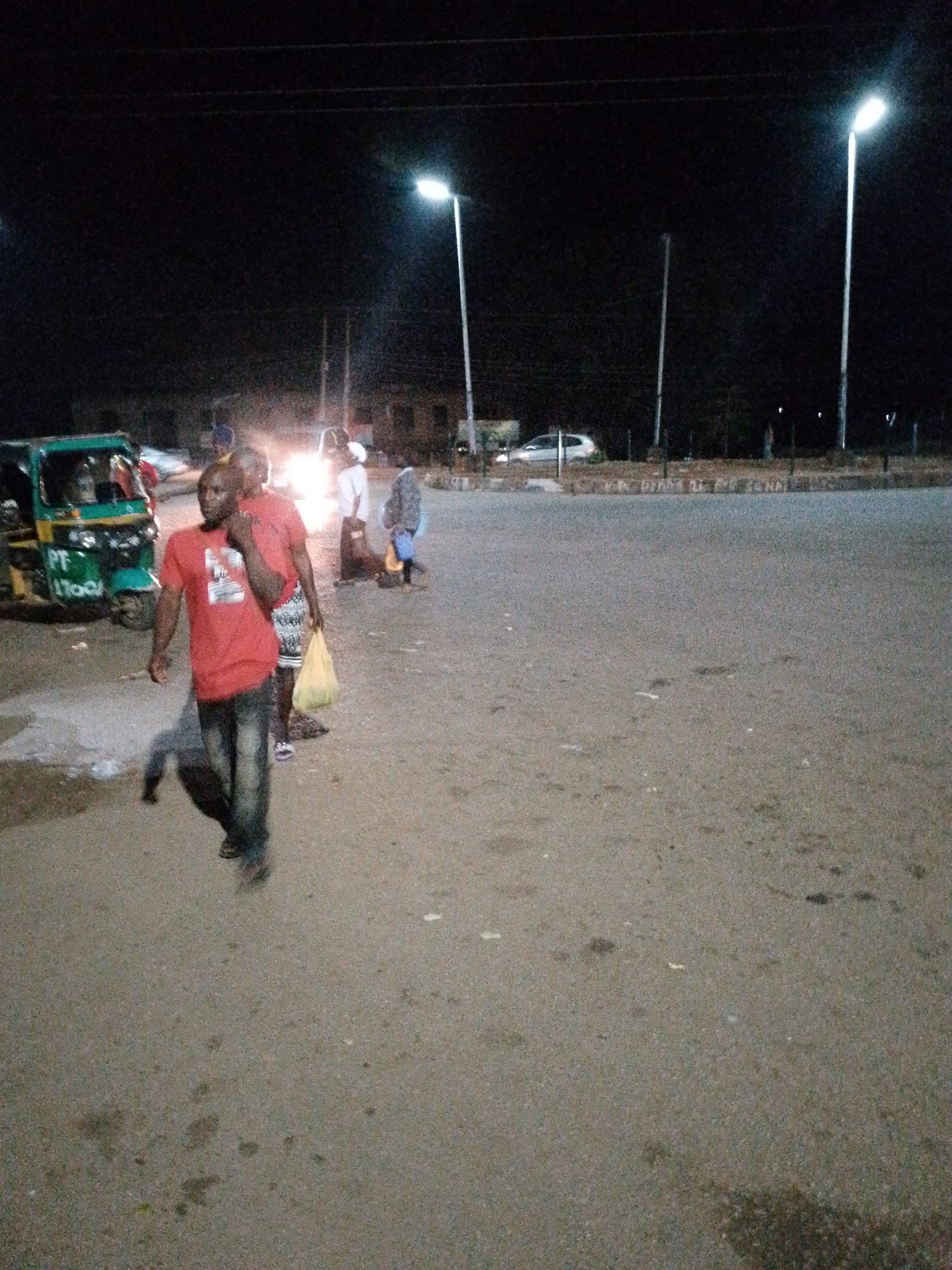 A low-light, slightly grainy, eye-level shot captures a street scene at night in an unidentified urban location. In the foreground, a Black man in a red t-shirt and dark jeans walks towards the left, his right hand raised to his chin. Behind him, another person, partially obscured and wearing a patterned skirt and a light-colored top, walks in the same direction, carrying a yellow plastic bag. To the left of the foreground figures is a three-wheeled green and yellow vehicle, possibly a tuk-tuk, with passengers inside. Further in the background, several streetlights illuminate the area. A white car is visible parked on the right side of the road. The ground is paved asphalt, with some scattered debris and a small puddle of water on the left. The overall impression is of a busy, but dimly lit, intersection or thoroughfare at night.