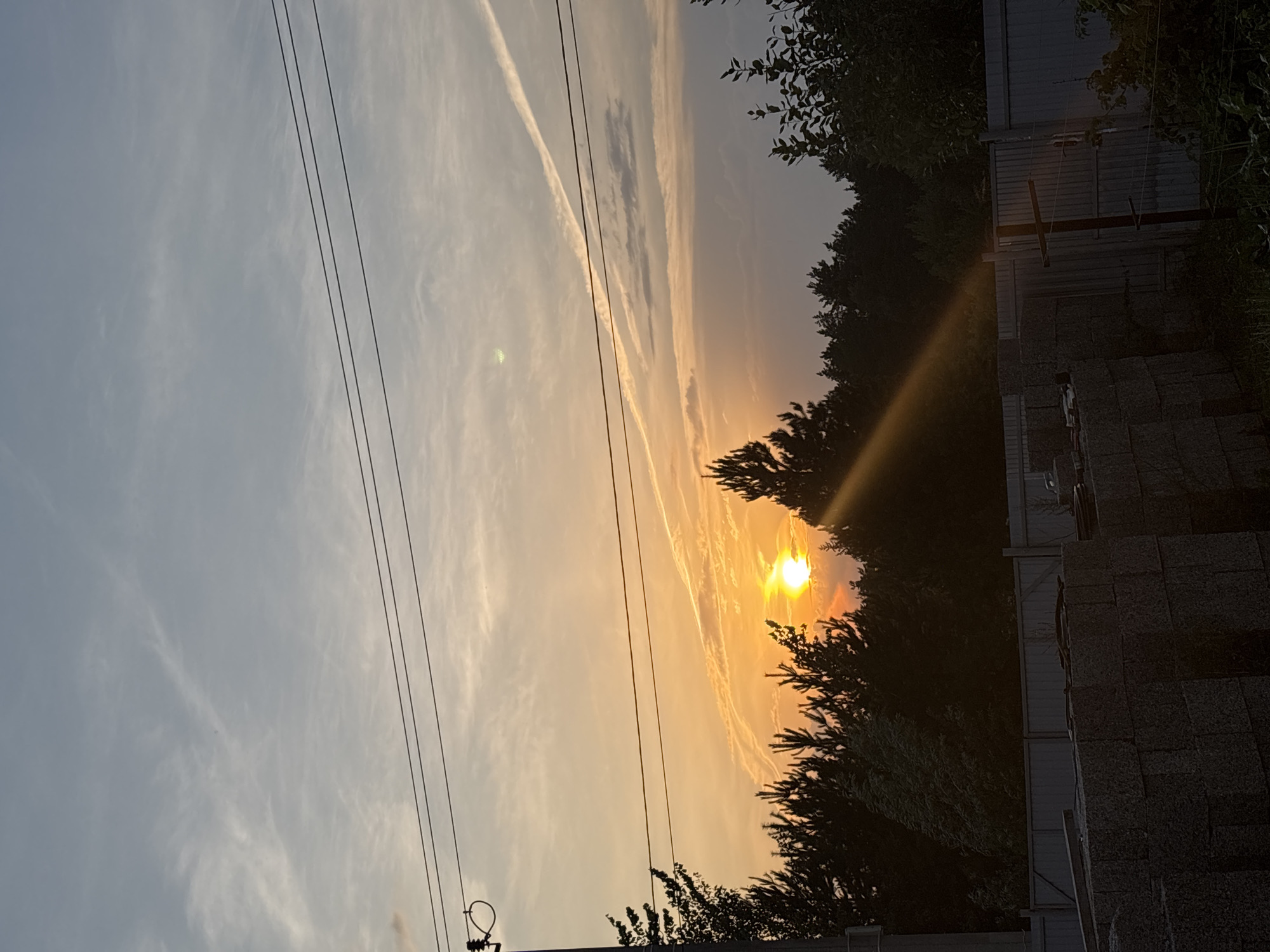 The image shows a sunset in a rural or suburban setting in Прикамский, Russia. The sun is a bright orange ball, low in the sky, casting a warm glow across the horizon. Wispy clouds stretch across the sky, some appearing as contrails from airplanes. In the foreground, silhouetted trees and power lines frame the scene. To the right, a fence and what appear to be stacked materials, possibly hay bales or construction materials, are visible. The overall mood is peaceful and serene, typical of a sunset. The time of day is clearly evening. There is no visible text in the image.