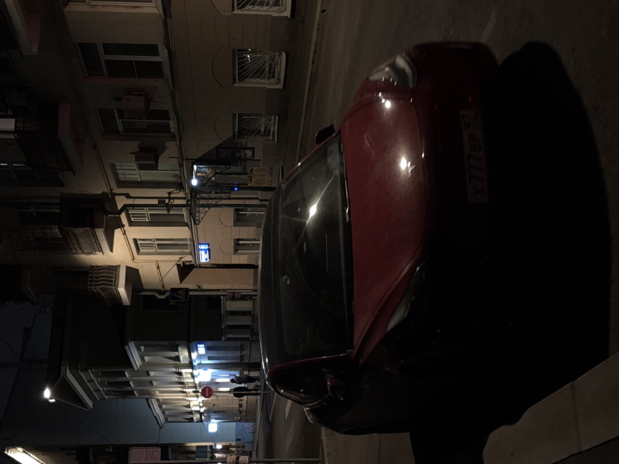 The image depicts a nighttime urban scene in Moscow, Russia. A red car is parked alongside a building. The license plate of the car reads "X777EP 77". Streetlights illuminate the facade of the building across the street, revealing multiple windows and architectural details. Pedestrians are present in the distance.