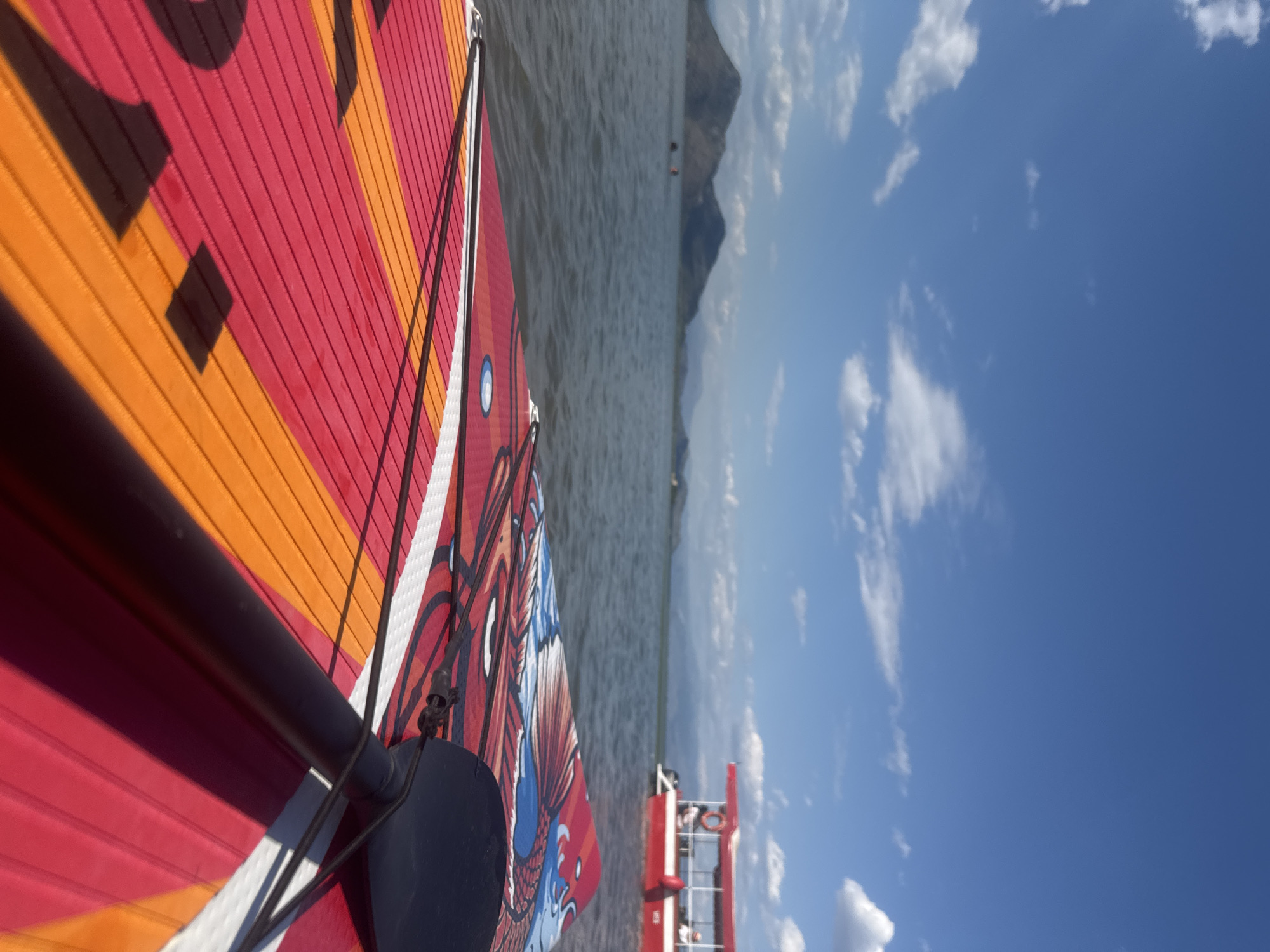 This is a first-person perspective shot from a paddleboard on a calm body of water, likely Lake Skadar near Seoca, Montenegro. In the foreground, a red and orange paddleboard with a graphic design is visible, along with a black paddle. In the middle ground, the water is dark and rippled, with a few people visible swimming or wading in the distance. To the right, a red and white boat with an open upper deck is cruising on the water. In the background, rolling, mountainous terrain is silhouetted against a bright blue sky dotted with white clouds. The lighting suggests it is daytime, likely midday or afternoon.