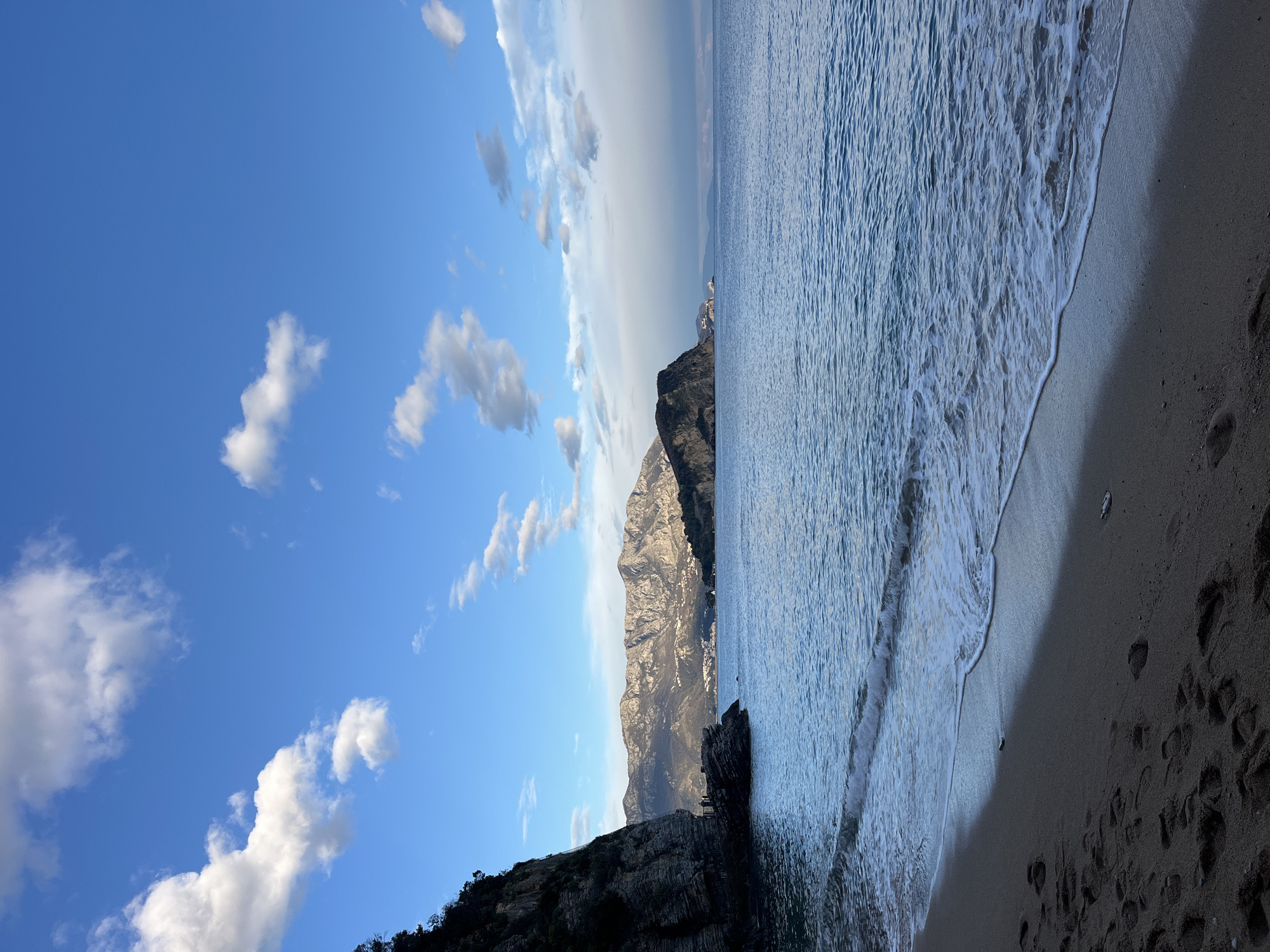 The image captures a coastal scene in Budva, Montenegro. A dramatic mountain range dominates the central view, with the Adriatic Sea to its right and a clear blue sky with scattered clouds above. The shoreline consists of a sandy beach, with waves gently lapping the shore. Footprints are visible in the sand. The photograph is oriented sideways.