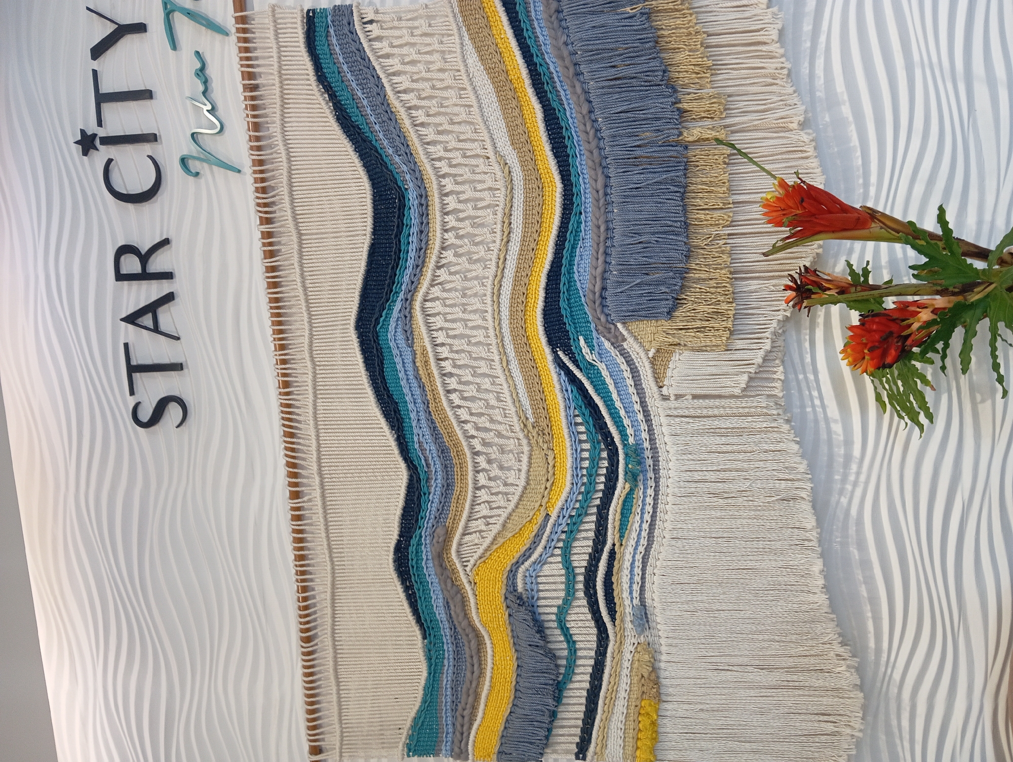 This is a photograph of a large, woven tapestry hanging on a wavy, white textured wall. The tapestry features a colorful, abstract design with horizontal bands of various textures and colors, including beige, yellow, gray, blue, and dark blue. It has a fringe at the bottom. Above the tapestry, the words "STAR CITY" are mounted in black, three-dimensional lettering, with a star symbol above the "A" in "STAR." To the right of "STAR CITY," the words "Nha Trang" are written in a turquoise, cursive font. The lower right portion of the image shows a bouquet of bright orange and red flowers with green leaves in a vase, partially obscuring the bottom of the tapestry. The overall impression is that of an interior decoration, possibly in a lobby or common area of a hotel or establishment in Nha Trang, Vietnam.