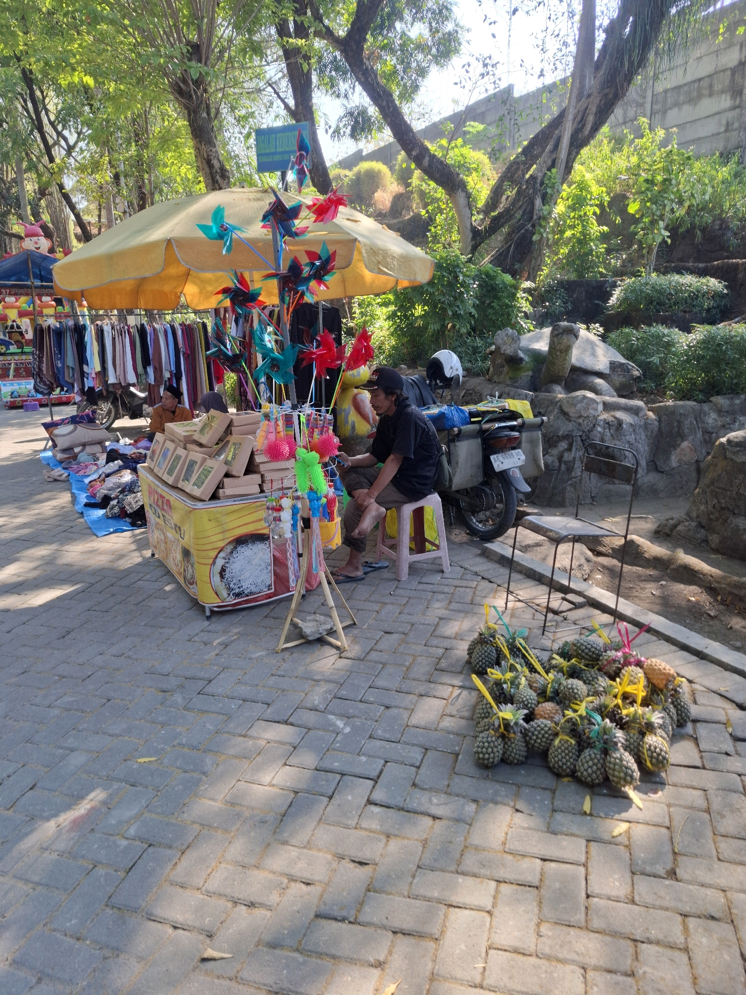 In Tawun, Indonesia, a vendor is selling goods from a stall set up on a paved area lined with trees. The stall features a yellow awning that covers racks of clothing and a display of pinwheels. Boxes are stacked on a yellow cart, and small toys are attached to a stand. In front of the stall, a man wearing a black shirt and a black cap sits on a pink stool. A motorcycle is parked behind the stall, and a metal chair is placed nearby. A pile of pineapples, tied with yellow and green ribbons, is arranged on the ground to the right of the vendor. The background consists of lush green foliage and rocky terrain, with a concrete wall visible further back.