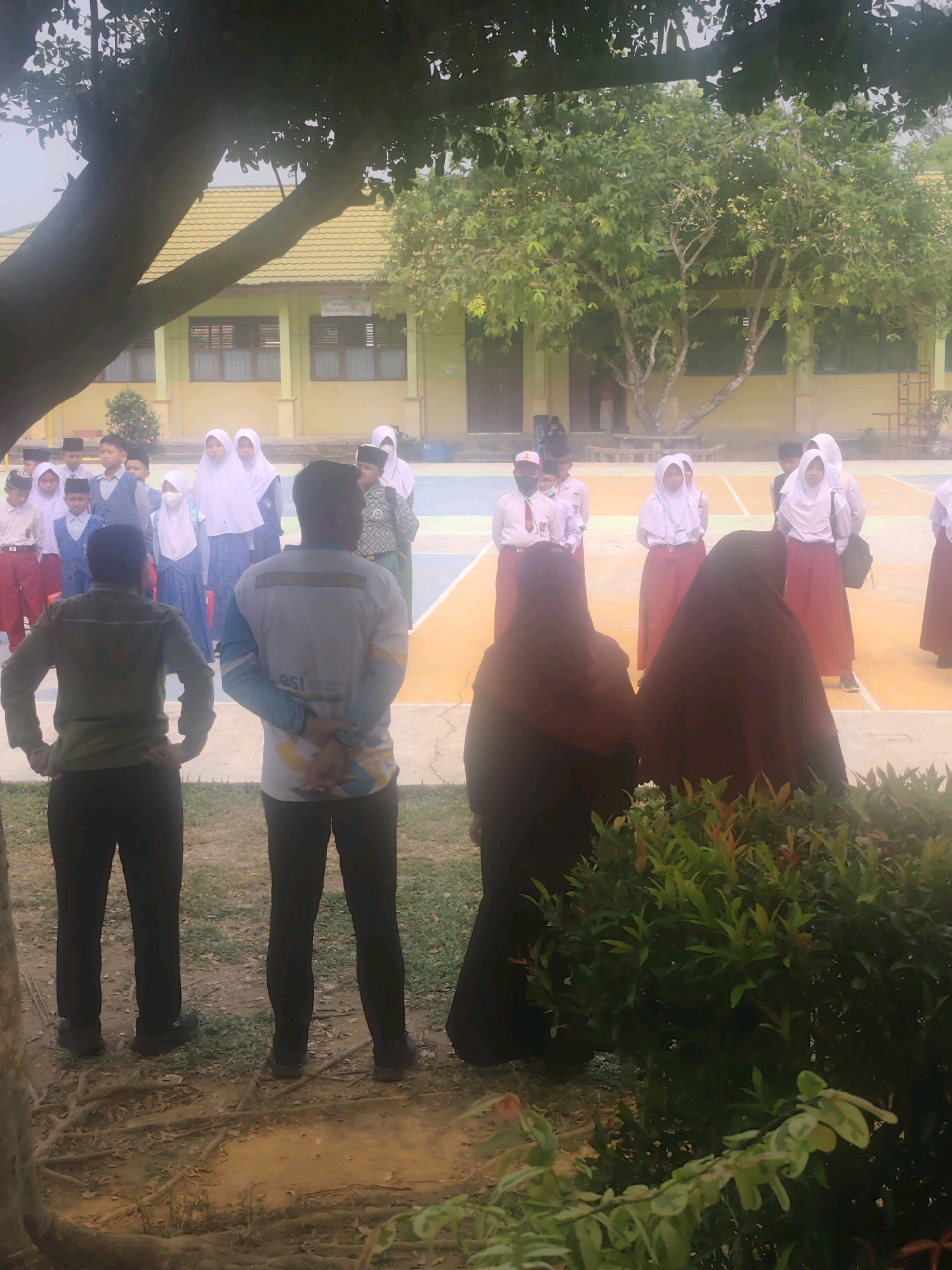 The image captures a scene outdoors, possibly on school grounds, in Pematang Reba, Indonesia. A group of students, some wearing white headscarves and red skirts, stand in formation on a colored court. Two adults, one wearing a khaki shirt and pants, the other a t-shirt with a graphic design and dark pants, face away from the camera. In the foreground, two individuals are partially obscured by a bush. Behind the students, a building with a yellow roof and cream-colored walls suggests an institutional setting. The environment includes a large tree in the left foreground, its branches extending over the scene, and vegetation in the right foreground. The overall lighting suggests it is daytime.