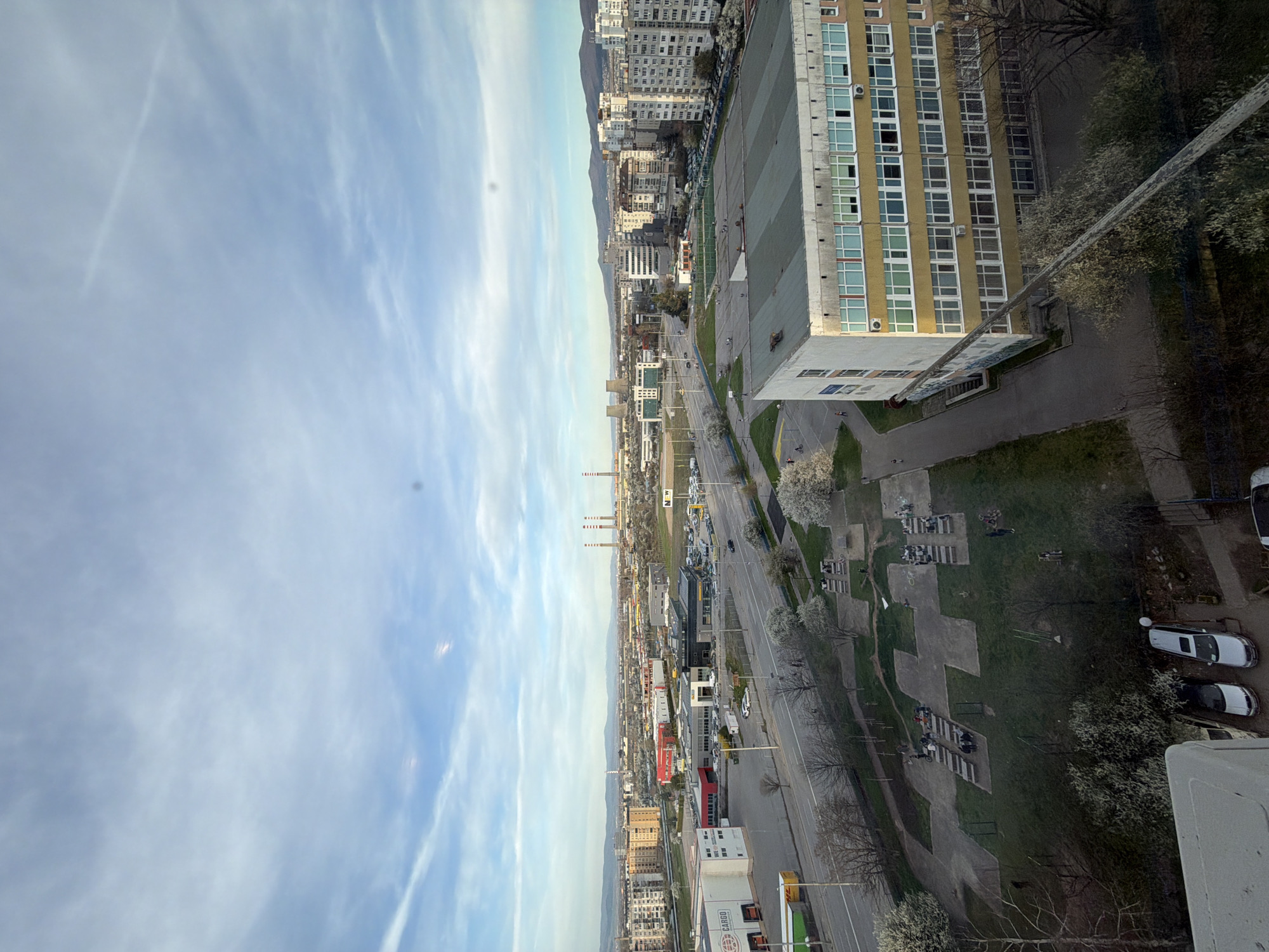 The image shows a high-angle view of a cityscape in Sofia, Bulgaria. The sky is pale blue with wispy clouds. Urban infrastructure includes apartment buildings and an adjacent multi-story building with a pale yellow facade. A large parking area sits next to it. A road runs through the city with light traffic. Farther down the road, there is a grouping of smokestacks. A small green space with trees, walkways, and concrete benches is adjacent to the yellow building. On the side of the image, there is a vertical concrete edge that suggests the vantage point of the camera is from inside a building. Several cars are parked alongside the concrete edge.