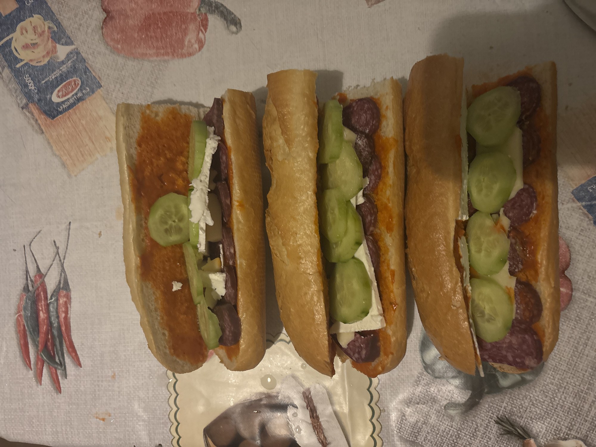 Three freshly prepared sandwiches, resembling sub-style baguettes, are laid out on a patterned tablecloth in an indoor setting. Each sandwich is cut lengthwise and generously filled. The base layer consists of a bright orange-red spread, likely a paprika-based relish such as lutenitsa or ajvar. Layered above this are numerous round slices of dark red salami or cured sausage. Interspersed among the meat slices are chunks or crumbles of white cheese, which could be feta or sirene, and several round slices of green cucumber, providing a fresh contrast.
The sandwiches are arranged in a row, slightly angled across the frame, with their open contents clearly visible. The bread appears to be a light golden-brown, suggesting it is fresh.
The tablecloth features several distinct patterns. In the upper left corner, a portion of a "BARILLA" pasta box is visible, specifically labeled "SPAGHETTINI n.3" and indicating "500g" weight. Towards the bottom left, an illustration of multiple red and dark chili peppers adorns the fabric. A scalloped edge of what appears to be a decorative placemat or plate is seen beneath the sandwiches on the right side, along with some indistinct patterns.
The scene suggests a casual meal preparation or serving, likely a light lunch or snack. The combination of salami, white cheese, cucumber, and a pepper-based spread is consistent with Balkan culinary traditions, aligning with the Zlatograd, Bulgaria location. No individuals are present in the image, and the lighting indicates an indoor environment.