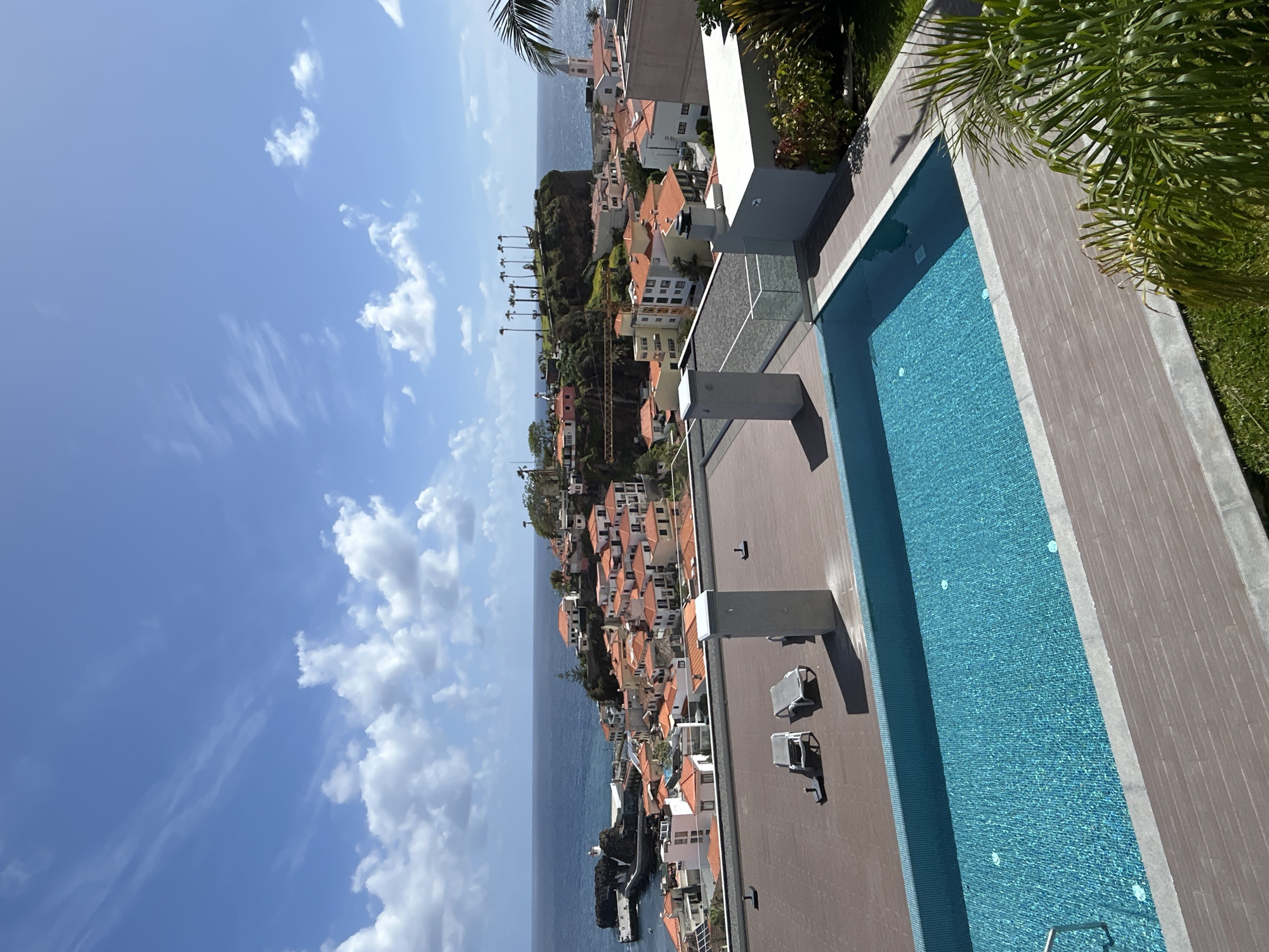 The image shows a view overlooking the coastal city of Câmara de Lobos, Portugal, featuring a swimming pool in the foreground. The pool has blue tiles, with a gray deck and lounge chairs nearby. The city's buildings, characterized by orange rooftops, are visible along the coastline. The sea is in the distance, and the sky is blue with white clouds. Palm trees are present in the image, one partially obscuring the view.