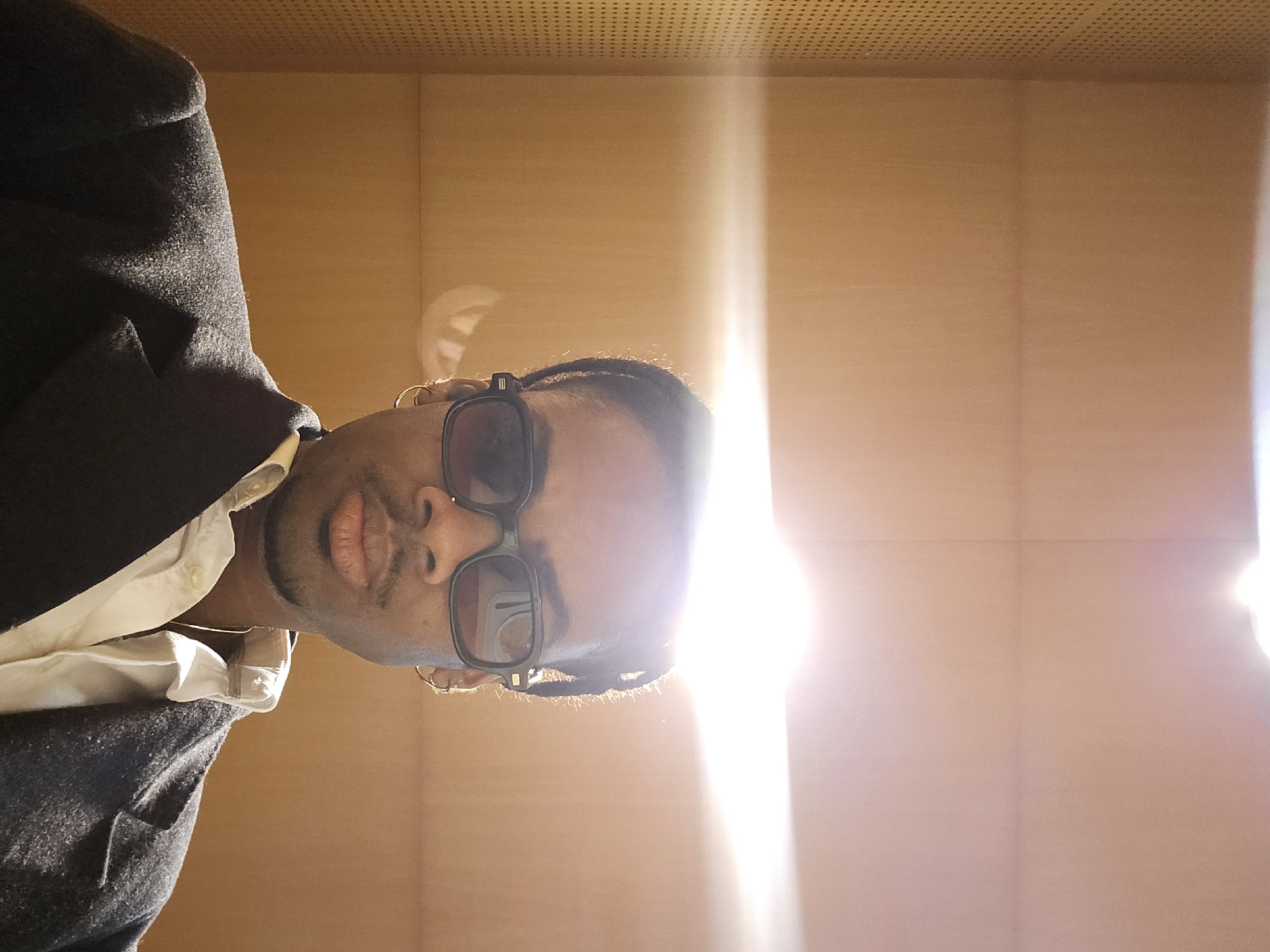 The image shows a person wearing glasses, a white shirt, and a dark blazer against a backdrop of light brown wooden paneling. The upper portion of the paneling has a grid of small holes. Bright light is streaming in, partially obscuring the person's face and parts of the background. The image is rotated counterclockwise. It is not possible to determine the specific location in Paris based on the image alone.