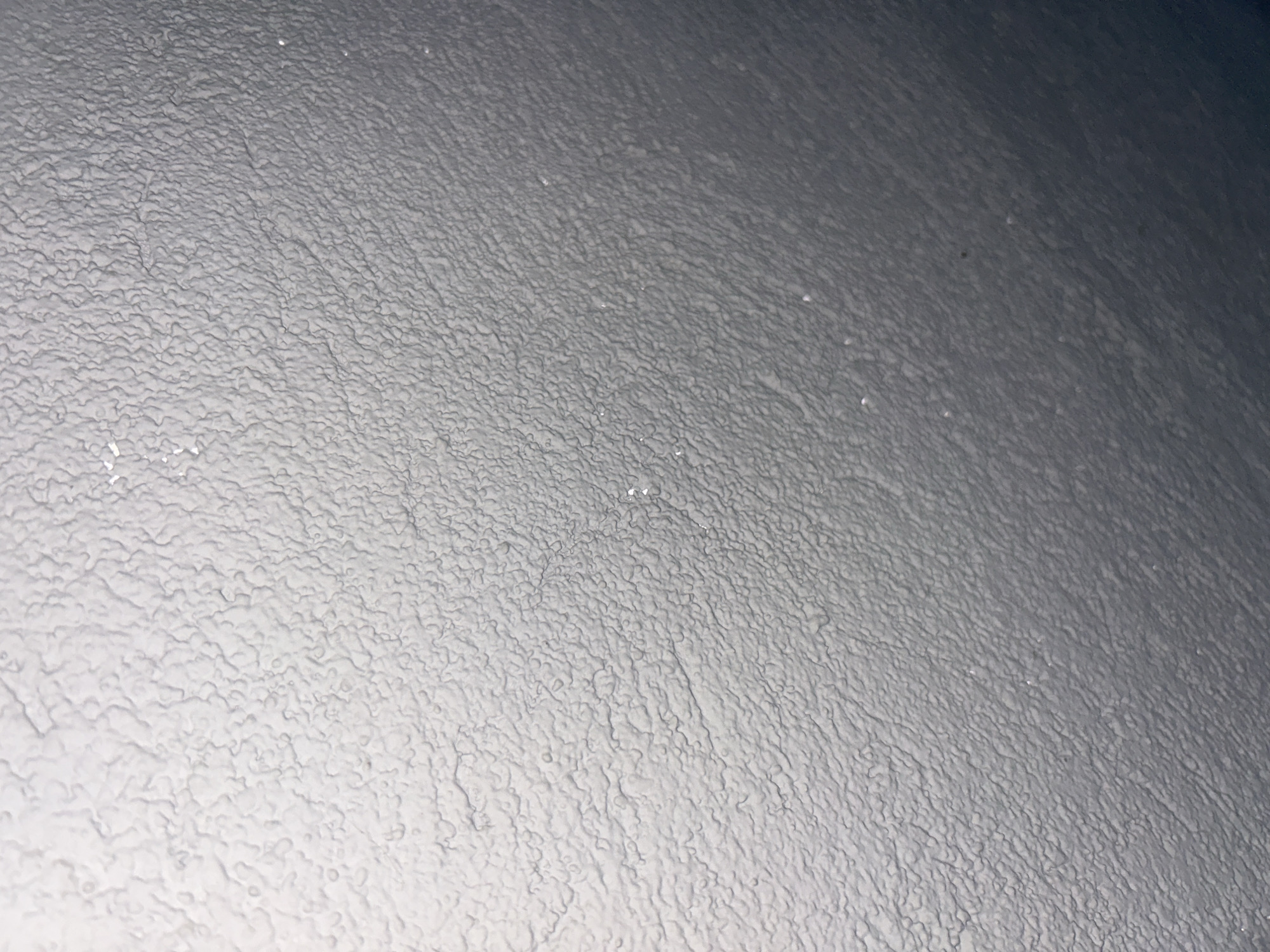 This media file presents a detailed close-up of a textured surface, primarily in shades of gray. The surface is covered with numerous small, irregular, and interconnected bumps and ridges, forming a granular or crystalline pattern. There is a clear diagonal light gradient across the image, transitioning from lighter gray tones in the lower-left to darker gray in the upper-right. Small, bright, reflective specks are visible across the textured surface, particularly in the lighter areas, suggesting the presence of ice crystals or frost. No identifiable objects, people, or text are present. The image focuses solely on the intricate details of the surface texture, which strongly suggests a natural formation like frost or ice covering an unseen underlying surface, indicative of cold weather conditions in a location such as Dunavtsi, Bulgaria. No specific activity or event is depicted.