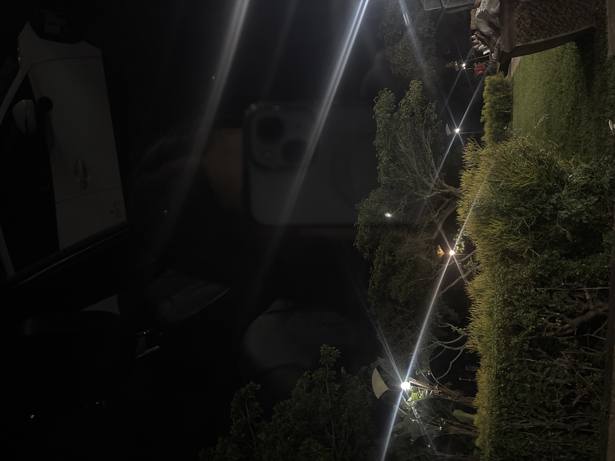 The image, taken at night, shows a car partially visible on the left, with foliage on the right side. Streetlights are scattered throughout the scene, creating streaks of light across the frame. The reflection of the photographer's phone is visible in the center. While specific locations are difficult to determine, based on the presence of lush foliage and street lighting, it would be plausible to place the location near a residential area or park in uMhlanga, South Africa.