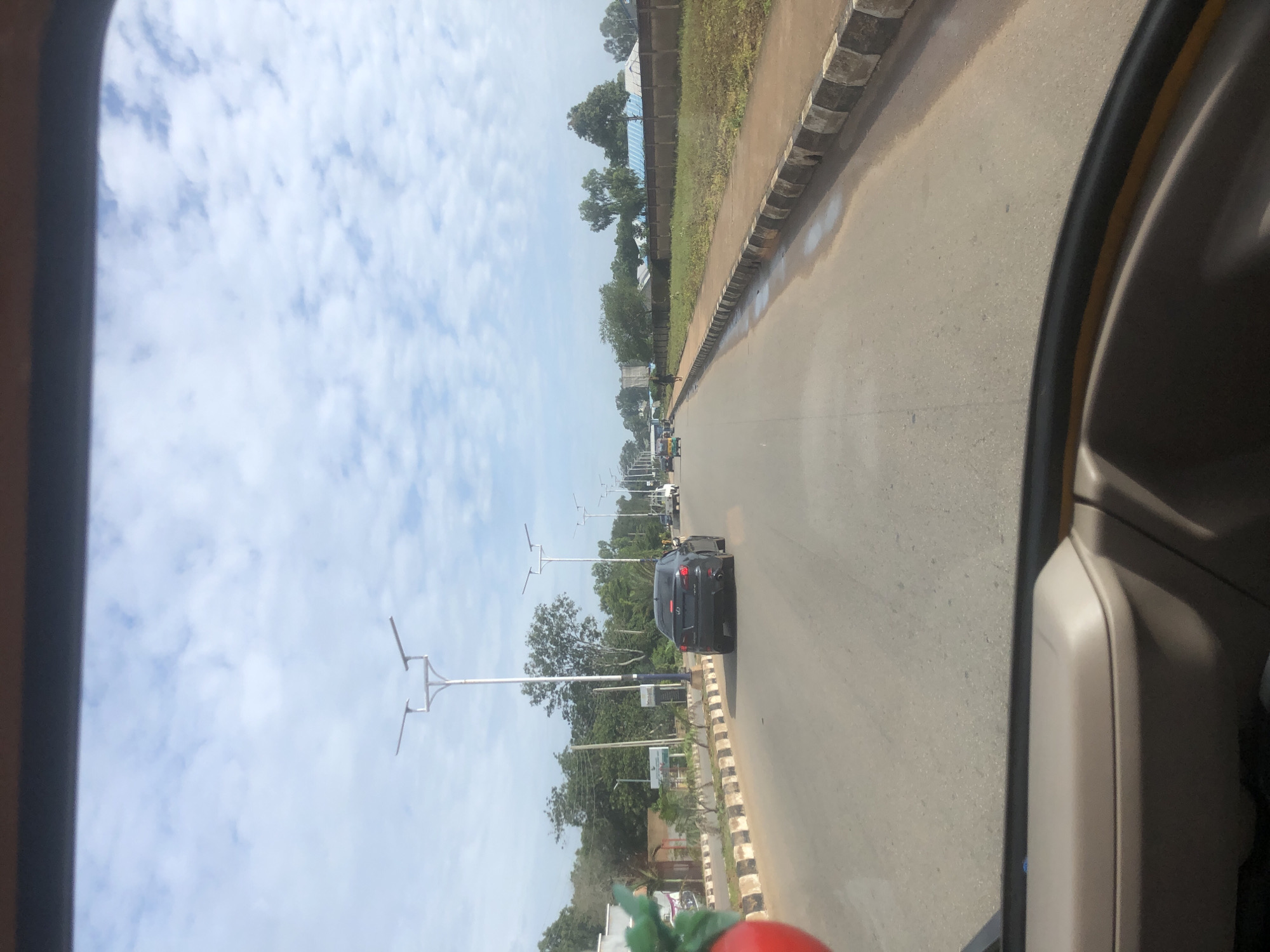 A black Lexus IS sedan travels down a paved road in Barakin Lerawi, Nigeria, under a partly cloudy sky. The vehicle is in the middle lane, with other cars visible further down the road. Streetlights line the sides of the road, and trees and buildings can be seen in the background. The road appears to be a main thoroughfare, with traffic signals and road markings indicating a well-maintained infrastructure. The scene suggests a typical day in a Nigerian city, with everyday activities taking place.