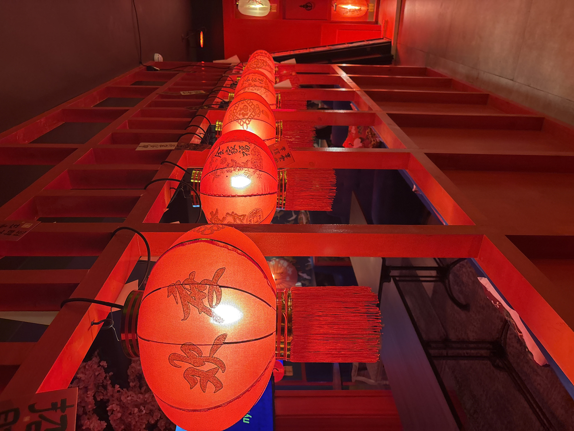 This image depicts the interior of a commercial establishment, likely a restaurant or bar, located in Saint Petersburg, Russia. The scene is characterized by a dominant red color scheme and Chinese-themed decor. A row of six illuminated red Chinese lanterns is suspended from a red wooden lattice partition that spans the midground.
The leftmost lantern prominently displays two large Chinese characters, "欢喜" (Huānxǐ), meaning "joy" or "delight," and features a dense red tassel fringe. The second visible lantern bears the Chinese characters "合家福" (Hé jiā fú), meaning "happiness for the whole family," also accompanied by red tassels. The subsequent four lanterns in the row are similarly red, illuminated from within, and feature various patterns or characters. Each lantern is suspended by a black cable attached to the upper horizontal beam of the wooden partition.
The red wooden lattice structure serves as a decorative divider, composed of evenly spaced rectangular vertical and horizontal beams. Small white paper slips with written text are affixed to several of these vertical elements. To the left of this partition, a seating area is partially visible, featuring dark tables and chairs. This area includes artificial pink cherry blossom decorations and a blue screen displaying partial text, including "ny." To the right of the partition, a clear walkway with a light-colored floor extends into the background, where additional red lighting fixtures and a white spherical lantern are visible. The ceiling above the entire space is dark. No individuals are present in the scene.