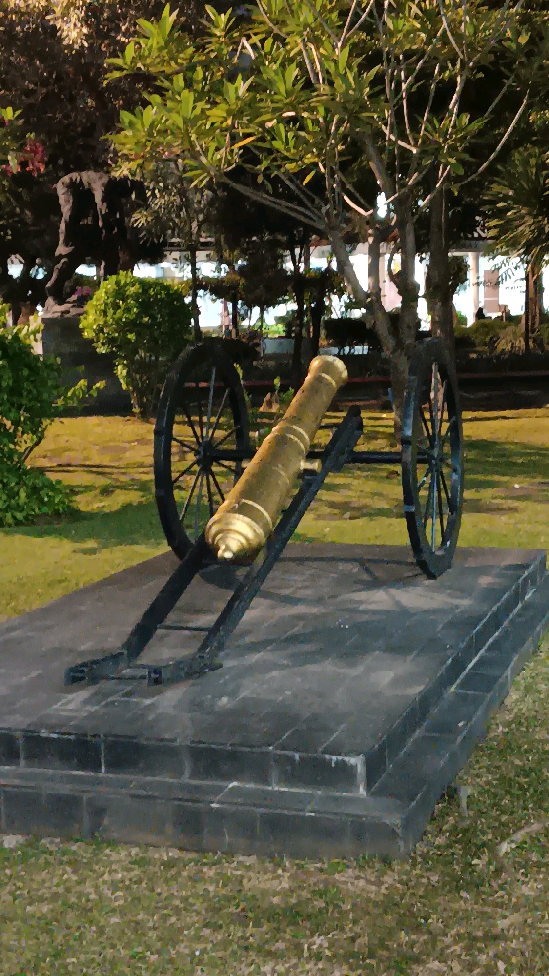 This image shows a cannon on a stone pedestal in a park-like setting. The cannon is brass-colored and mounted on a black wheeled carriage. The pedestal is made of dark gray stone blocks, with several tiers. In the background, there are trees with green leaves, and some buildings. The ground is covered with green grass and dry leaves. This appears to be an outdoor exhibit or historical monument in a public space. The specific location in Central Jakarta, Indonesia is not identifiable from the image alone.